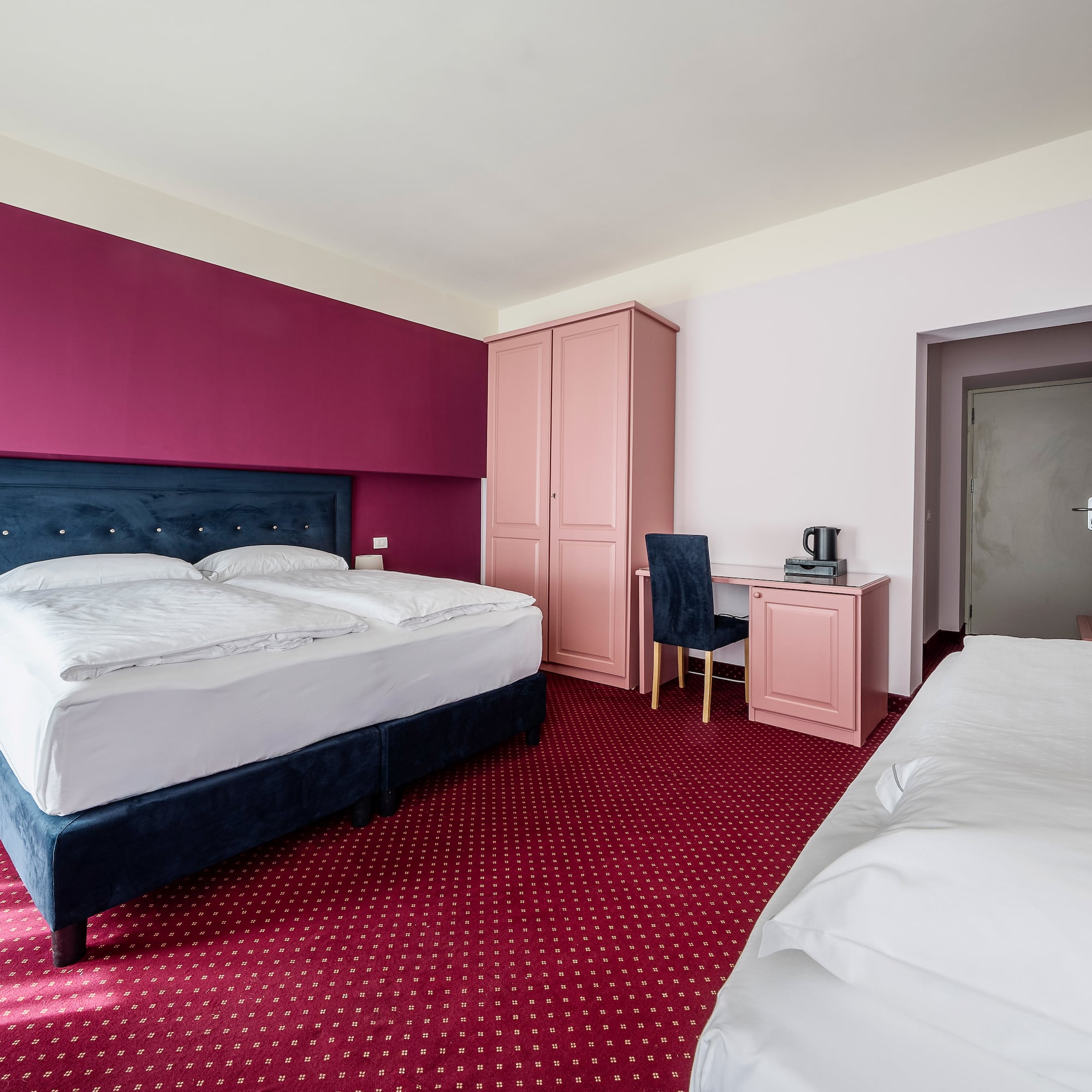 Hotel room with magenta walls, blue bed, red carpet, pink furniture, and extra bed.