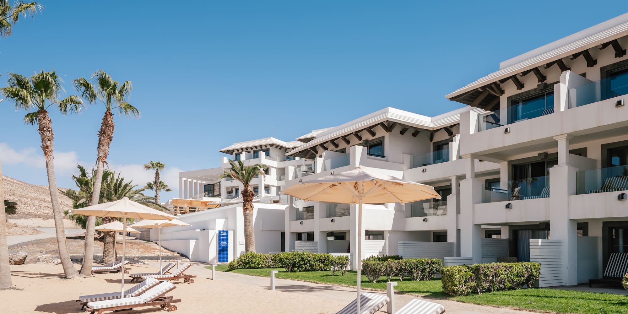 Sunny beach resort with white hotel buildings, palm trees, lounge chairs.