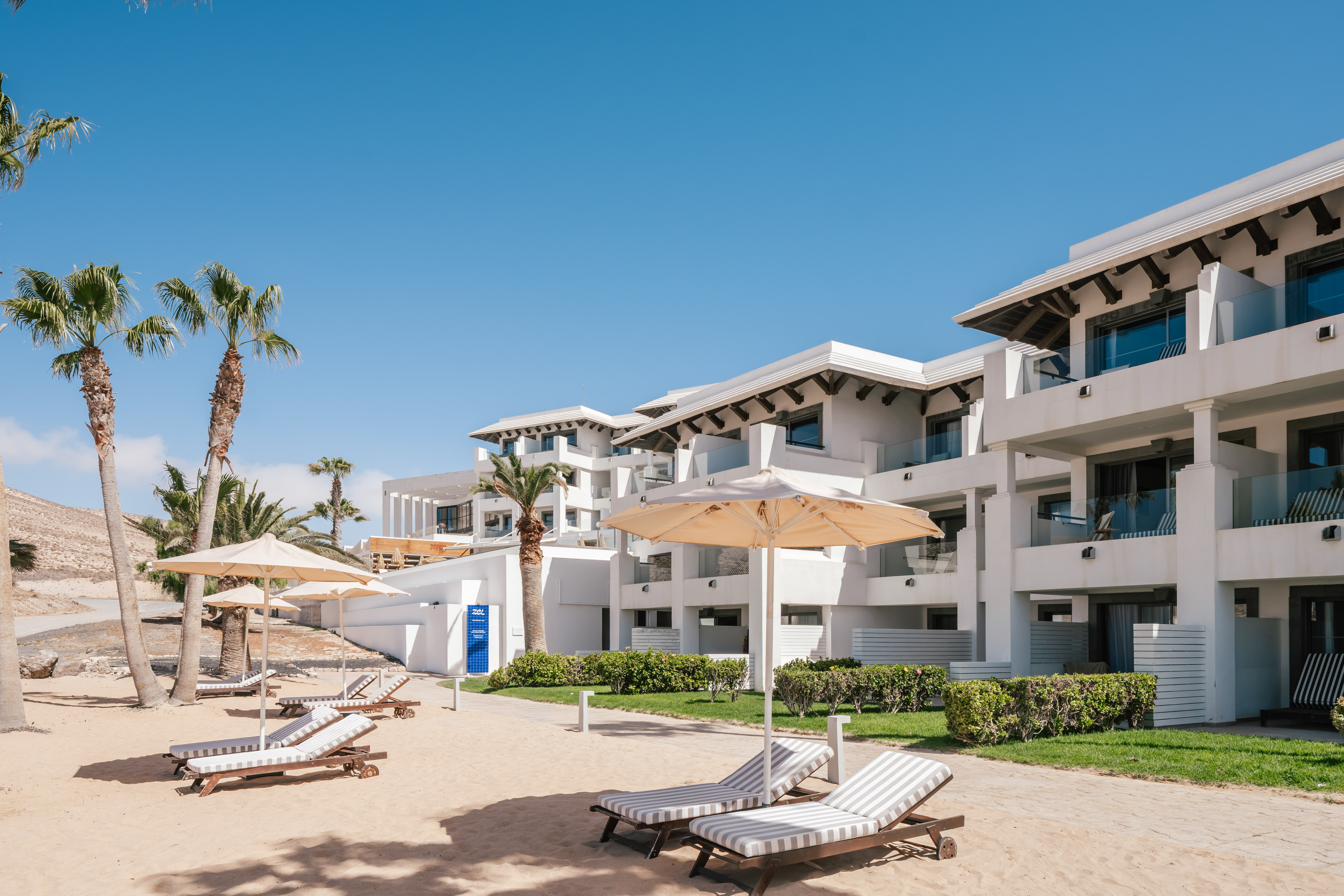Sunny beach resort with white hotel buildings, palm trees, lounge chairs.