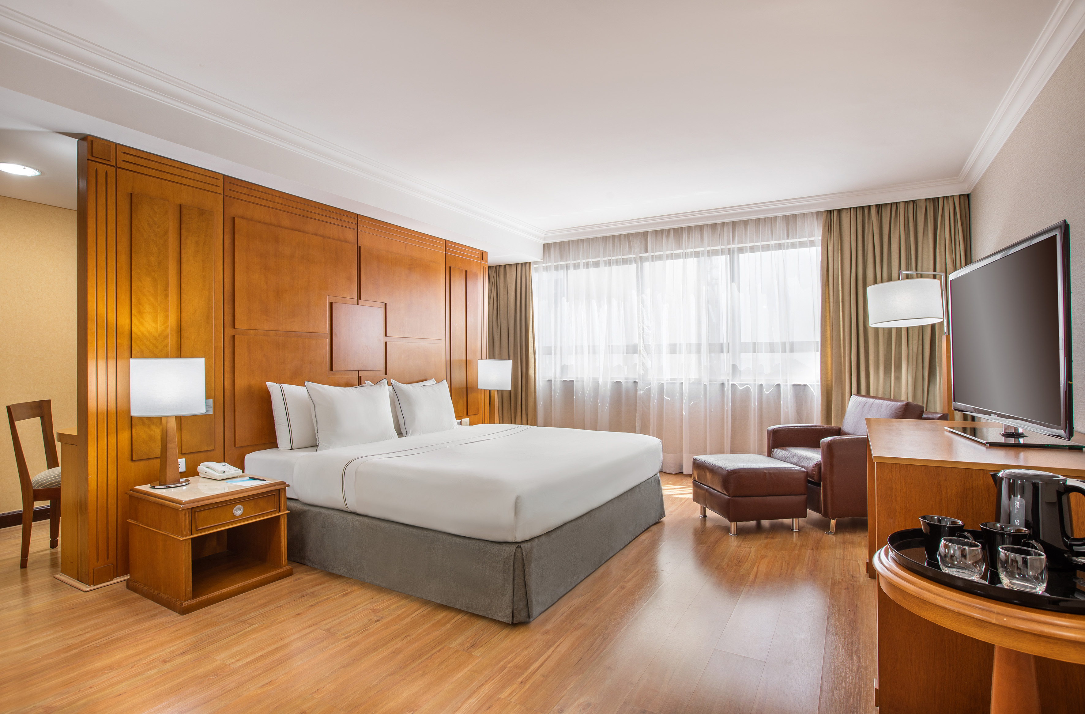 Hotel room with king bed, wooden wall panels, armchair, and bright window.