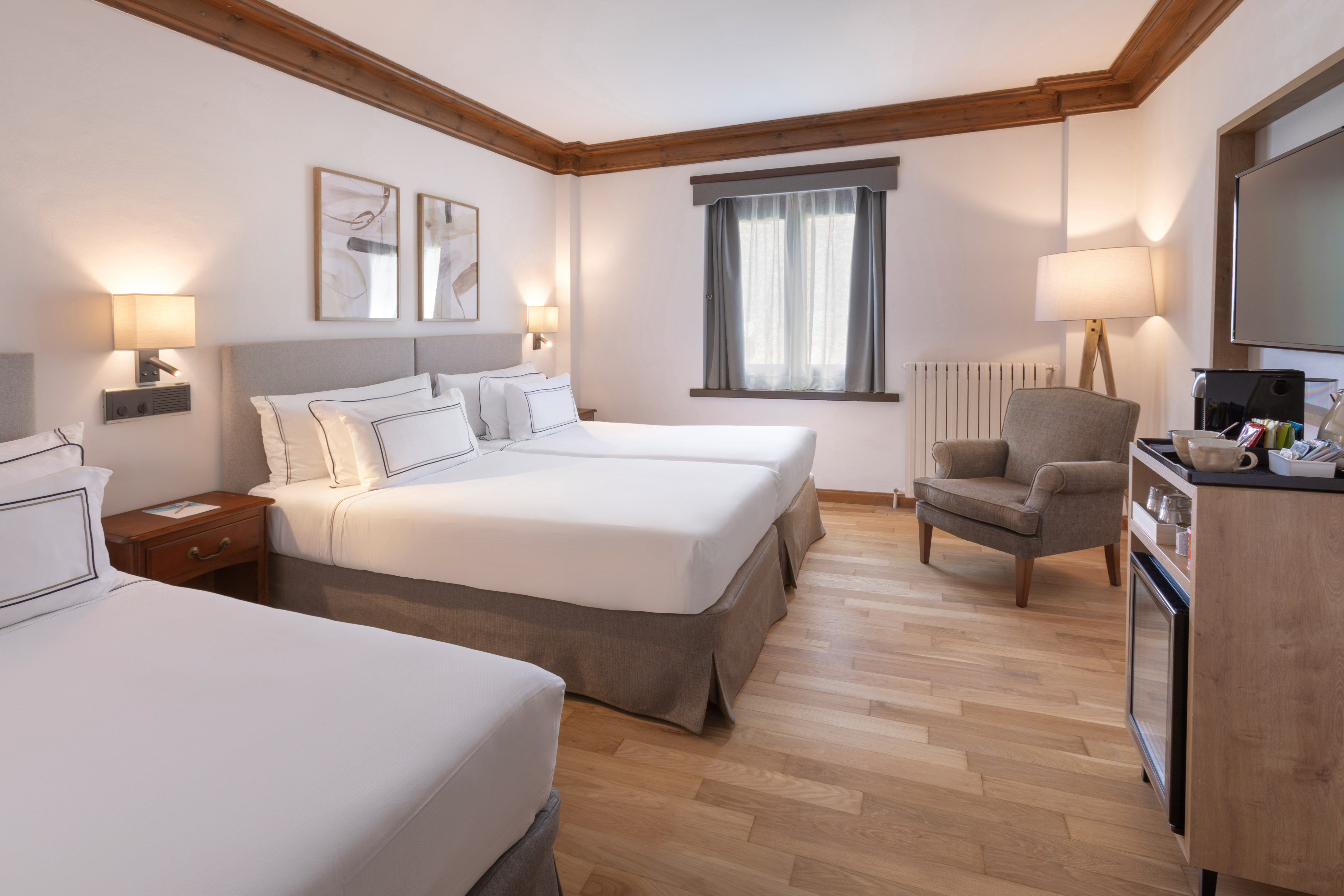 Hotel room with three white beds, wooden decor, armchair, and coffee setup.