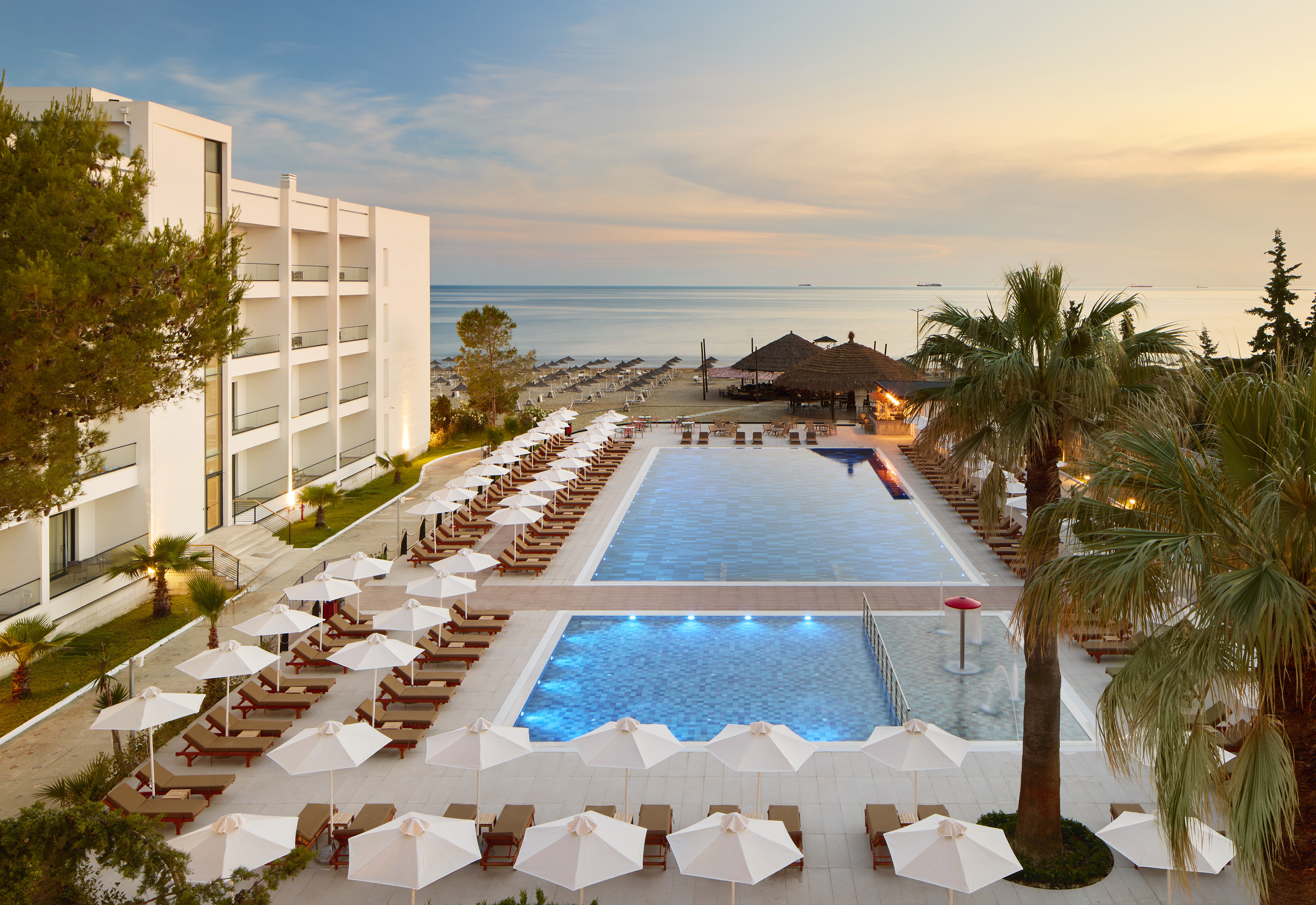 Resort with illuminated pools, sun loungers, beach, and ocean at sunset.