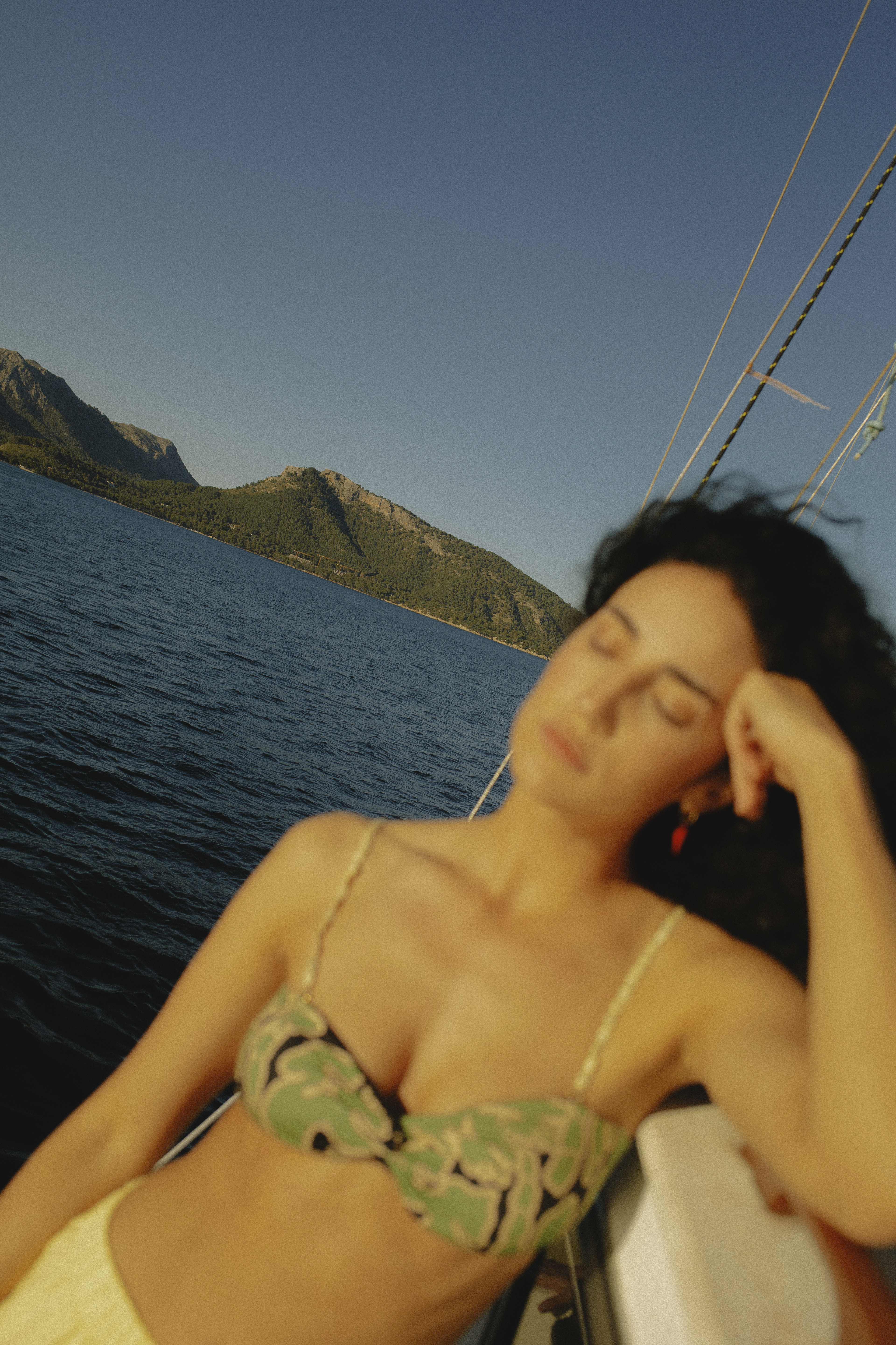 Relaxed woman in patterned bikini on boat, blue sea, and green mountains.