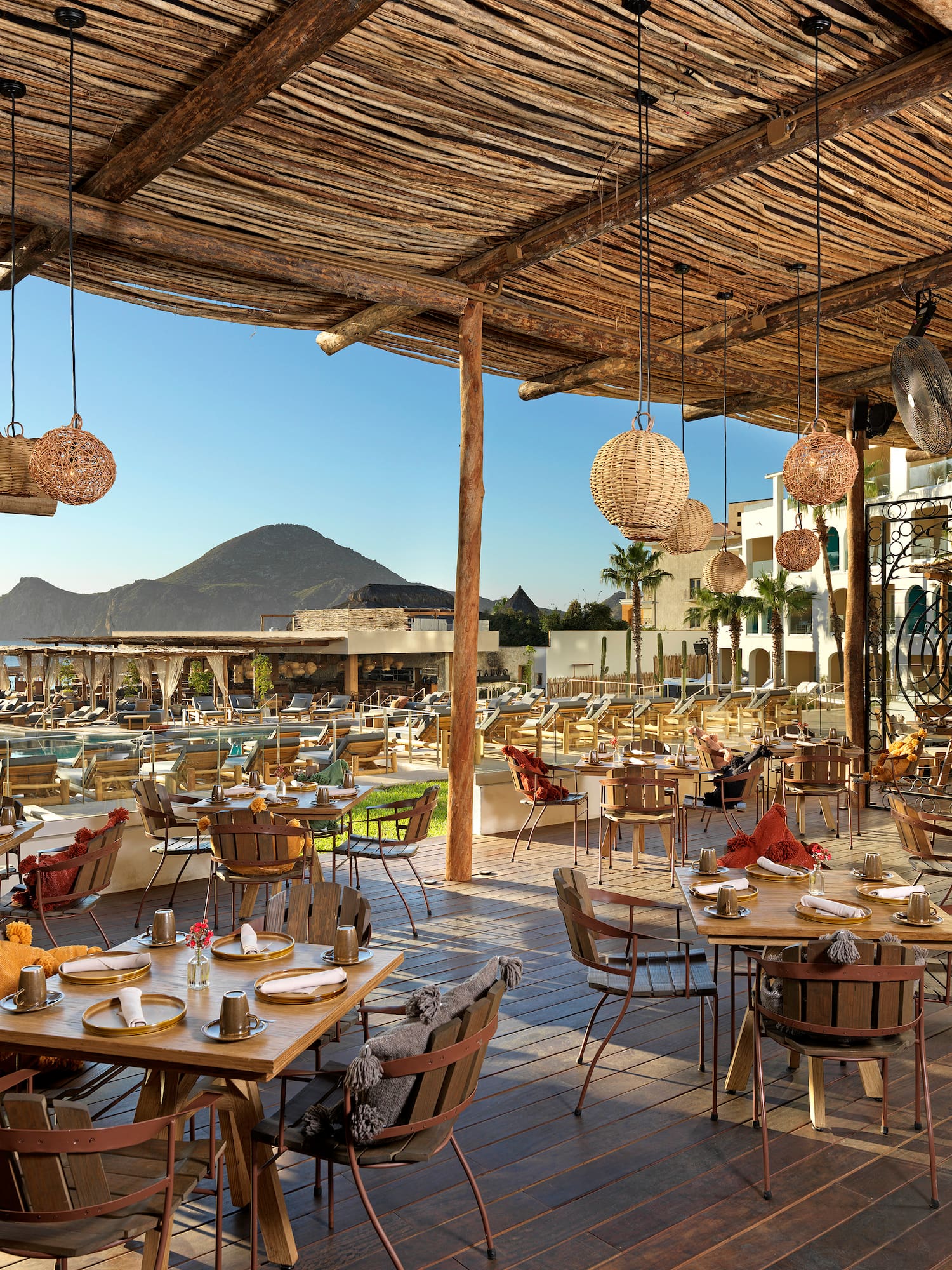 Sunny outdoor restaurant, woven roof, set tables, resort pool, ocean, mountains.