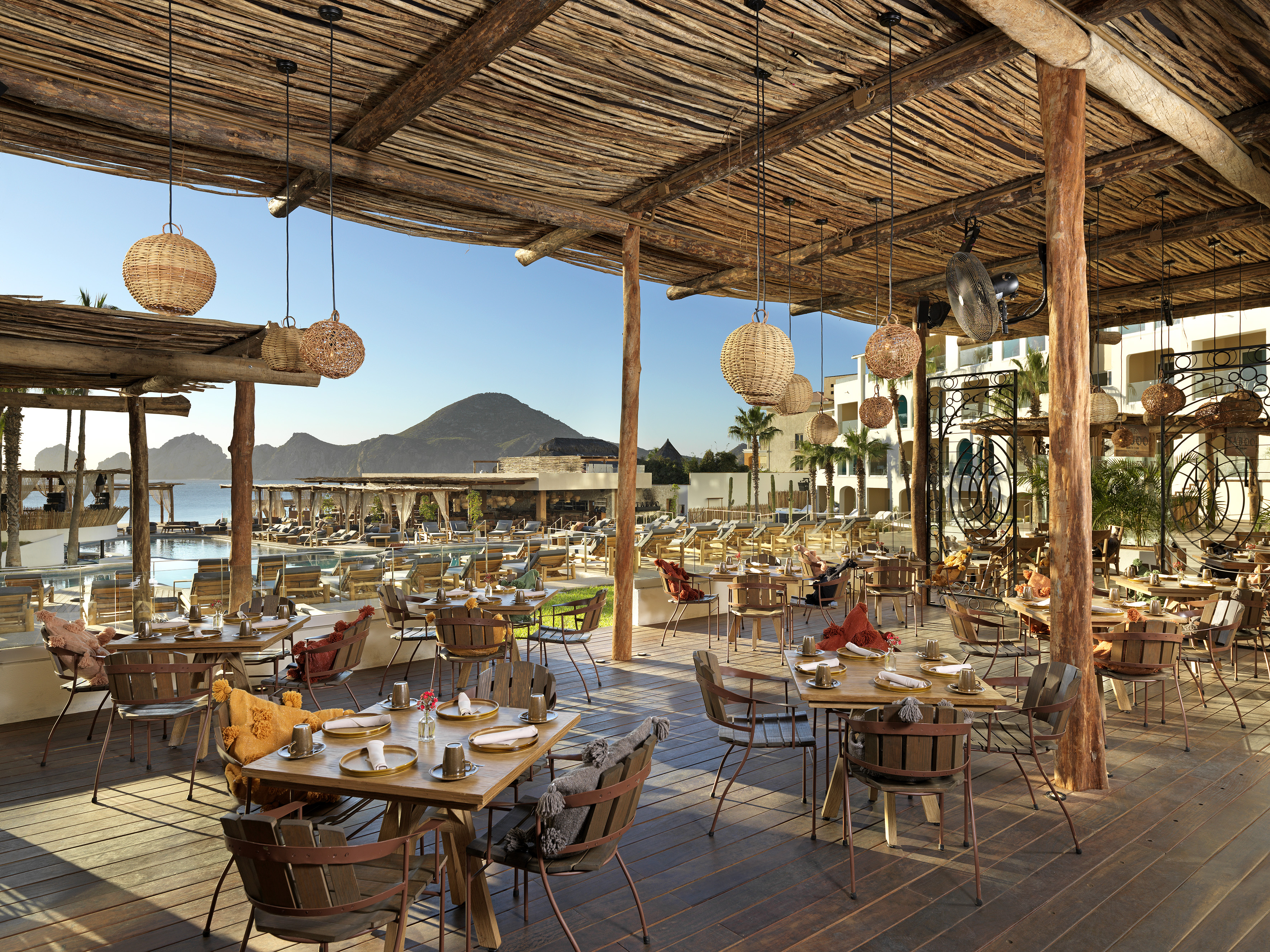 Sunny outdoor restaurant, woven roof, set tables, resort pool, ocean, mountains.