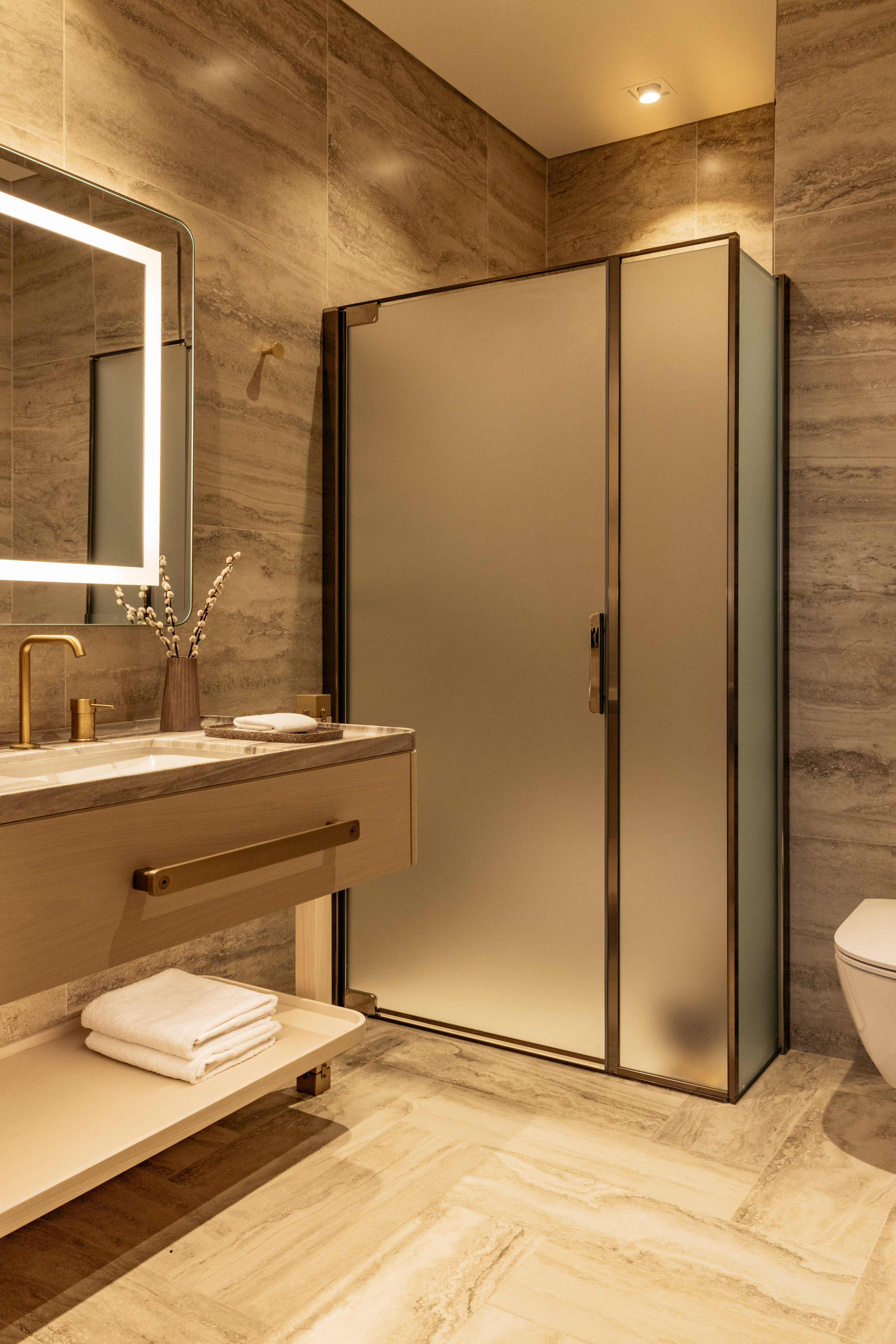 Modern bathroom: wood vanity, gold fixtures, backlit mirror, frosted shower.