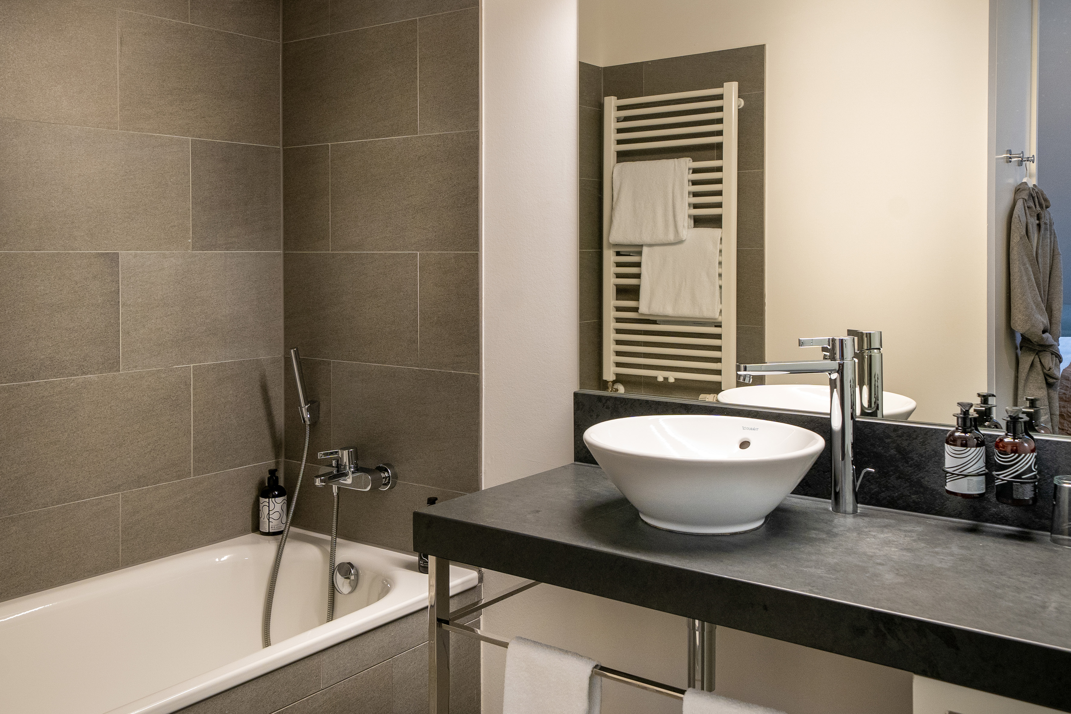 Modern hotel bathroom with gray tiles, bathtub, vessel sink, and towel warmer.