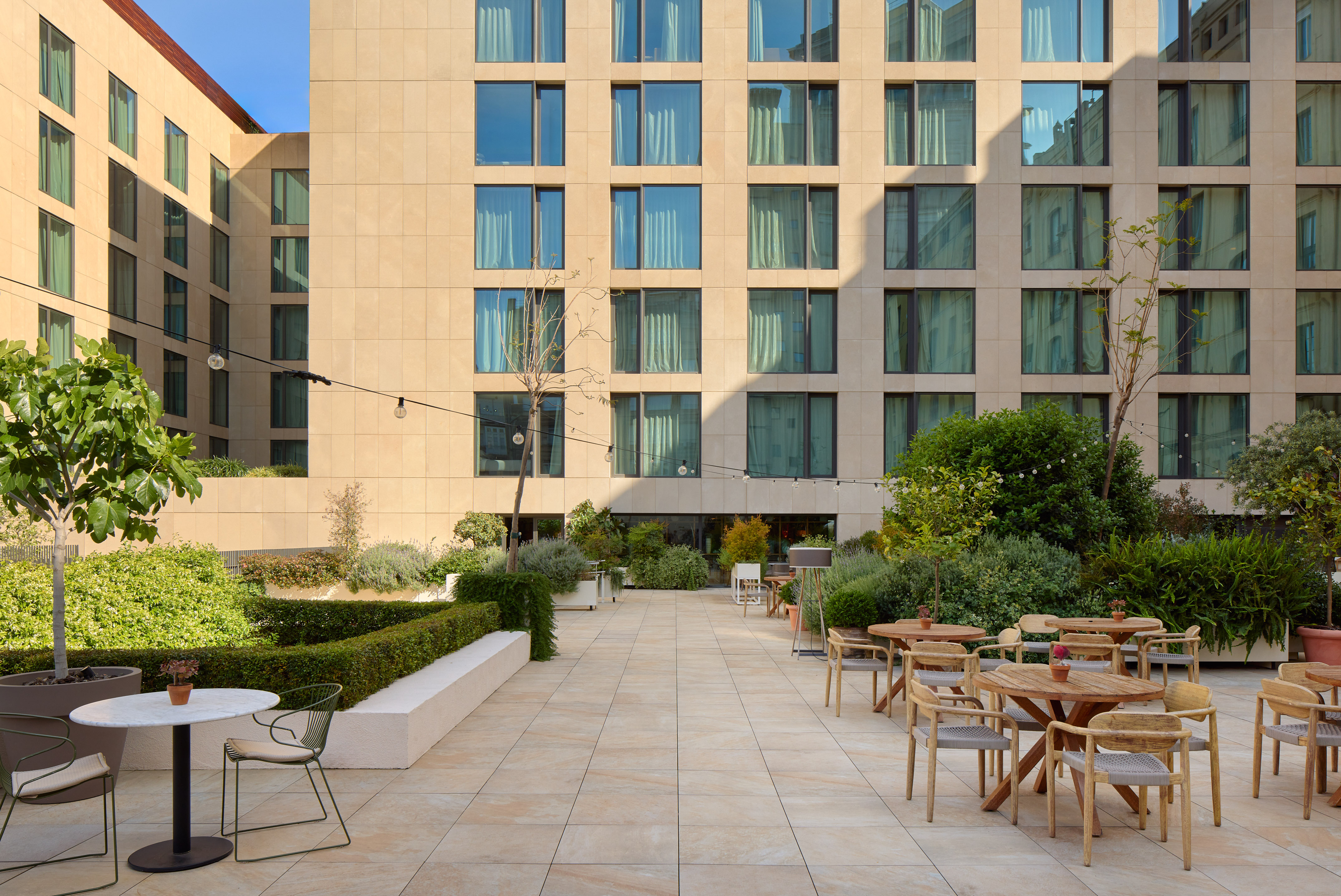Hotel outdoor terrace with dining tables, green plants, and contemporary building.