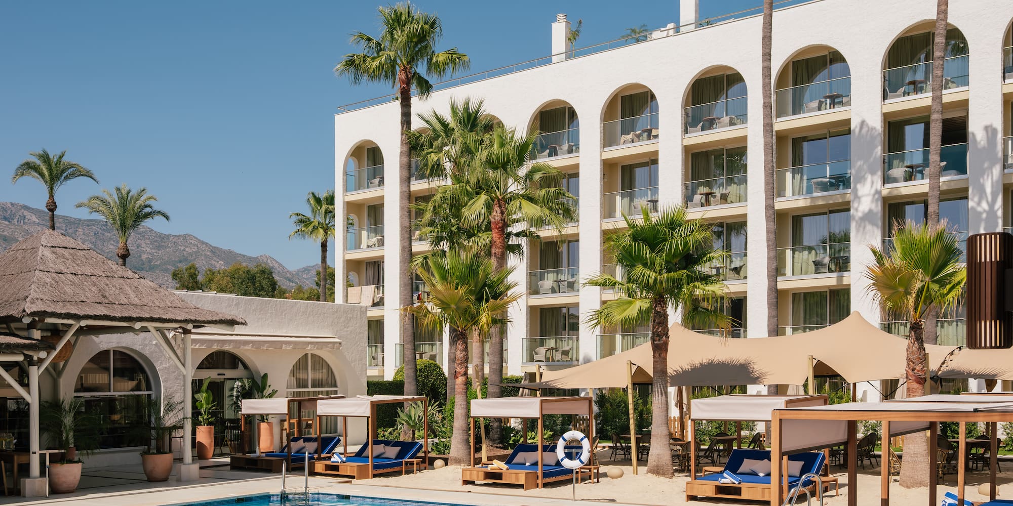 Luxury hotel pool with palm trees, cabanas, and white arched building.