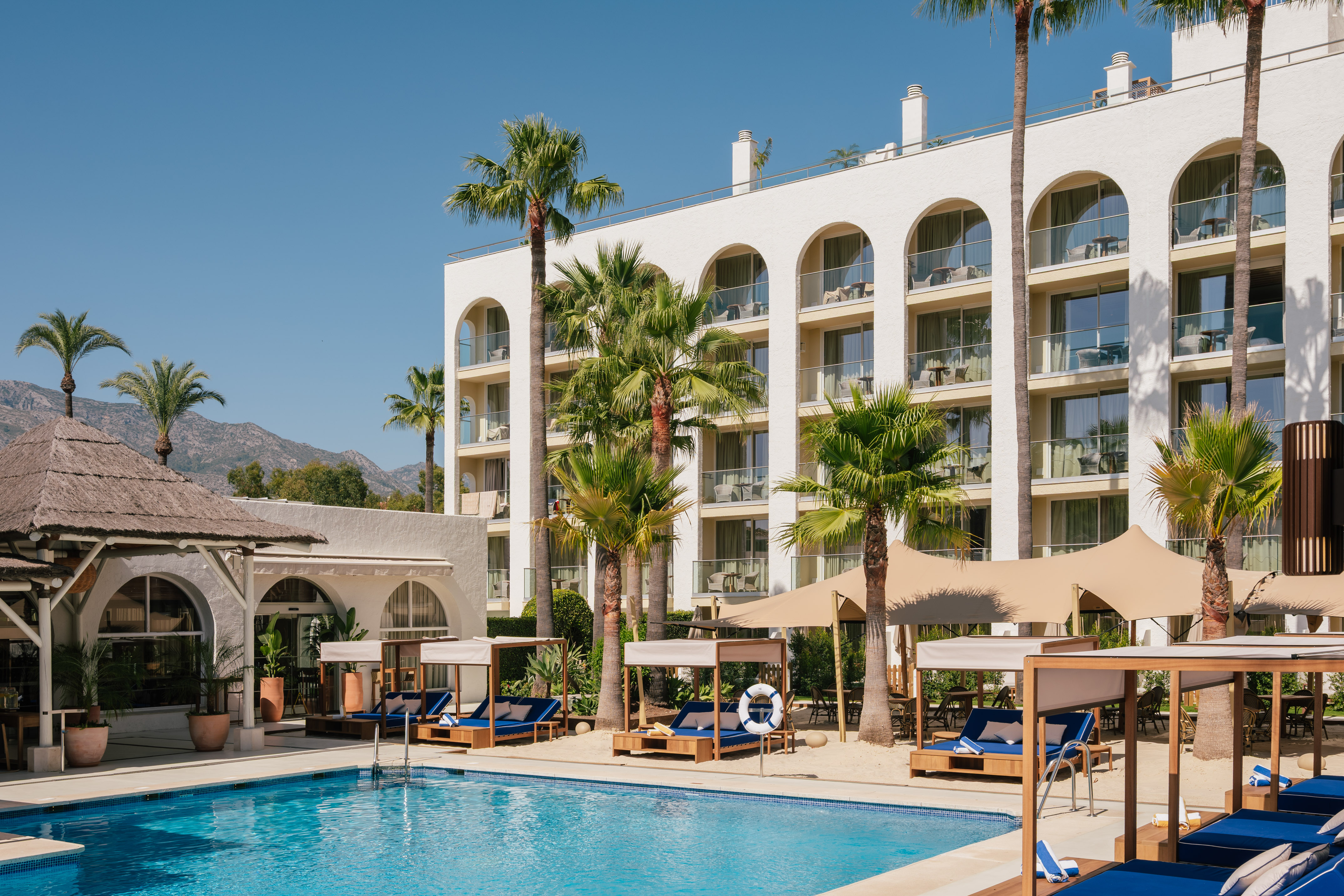 Luxury hotel pool with palm trees, cabanas, and white arched building.