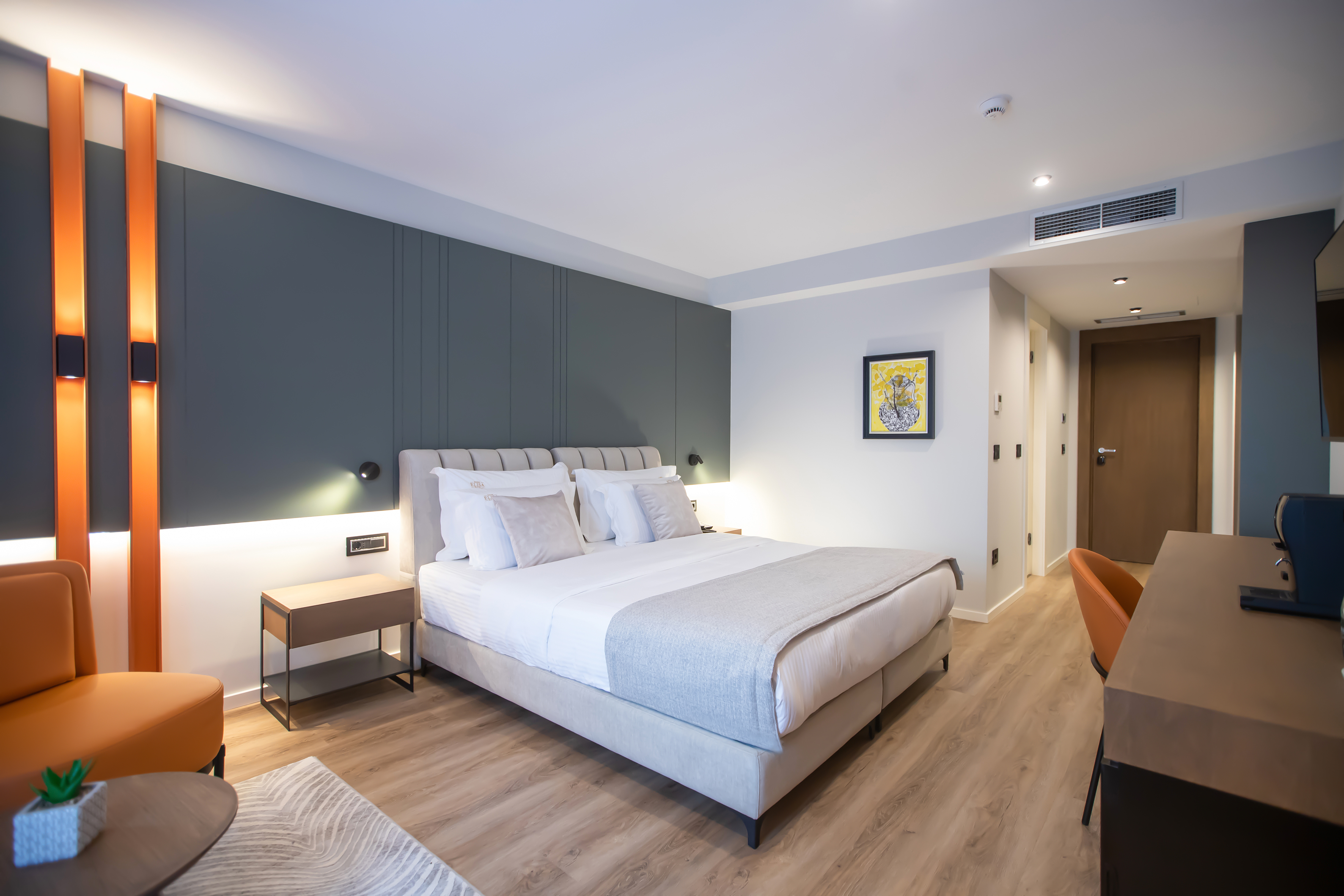 Modern hotel bedroom with bed, orange chair, dark grey wall, and wooden floor.
