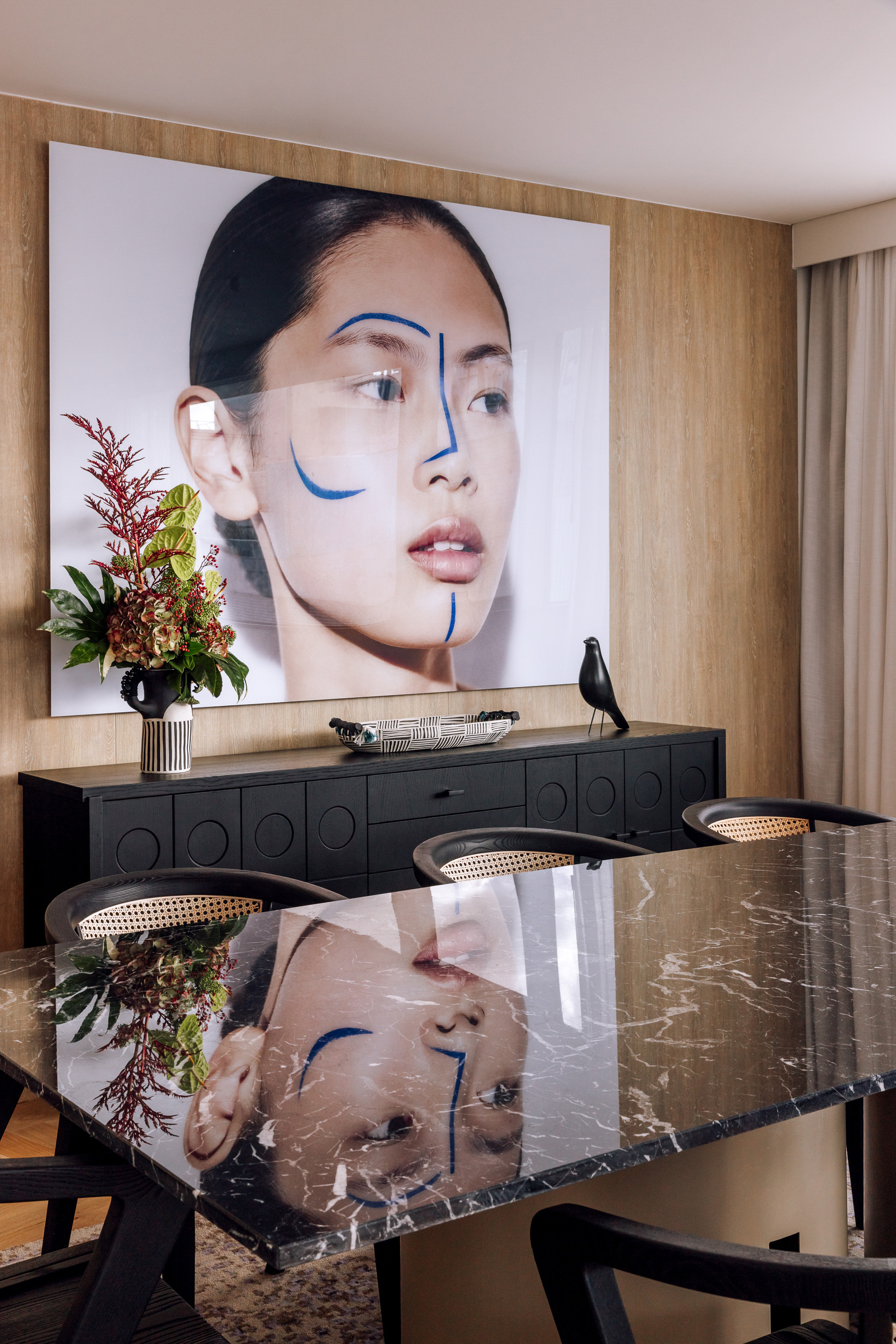 Chic dining room: oversized abstract face art, black credenza, marble table, cane chairs.