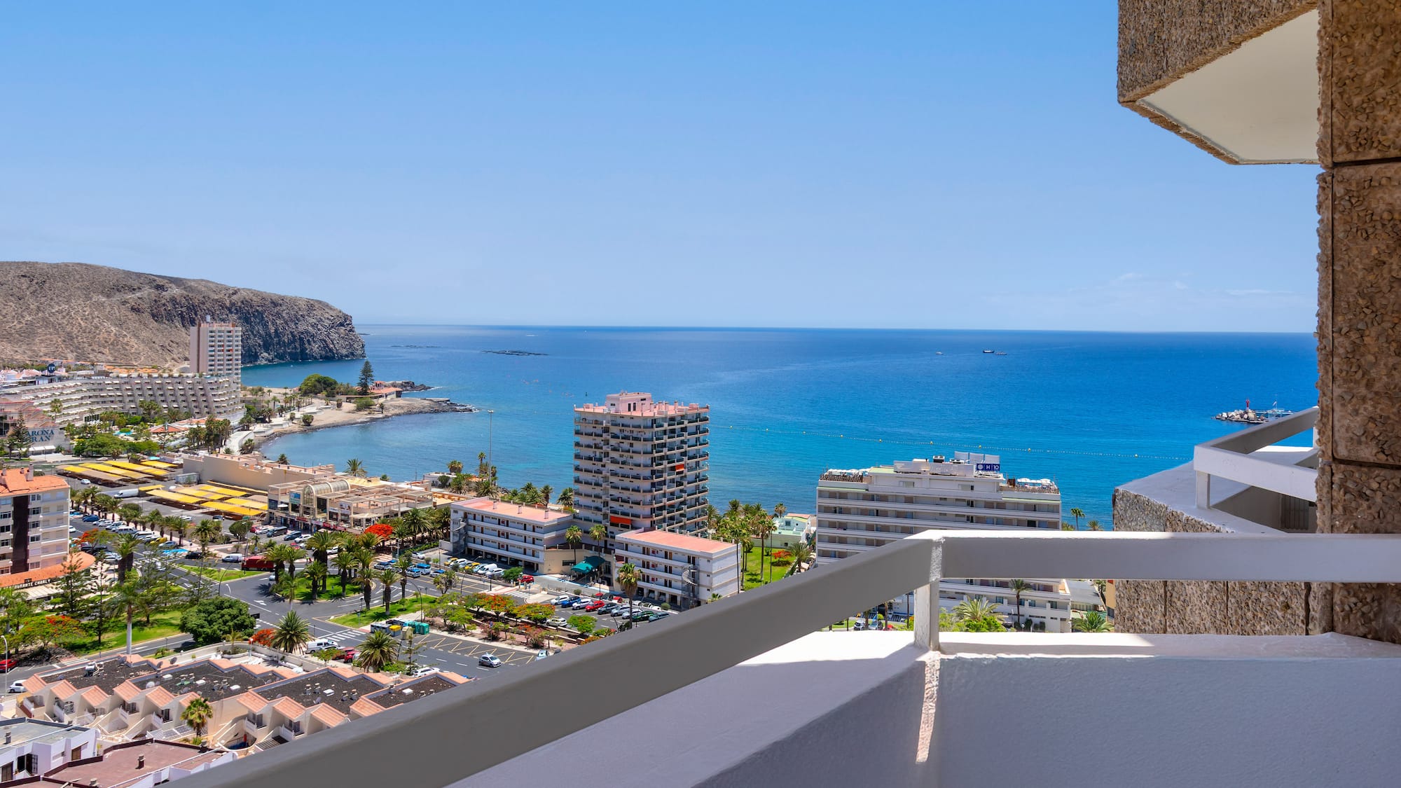 Panoramic balcony view of a sunny coastal town, blue ocean, and rugged cliffs.