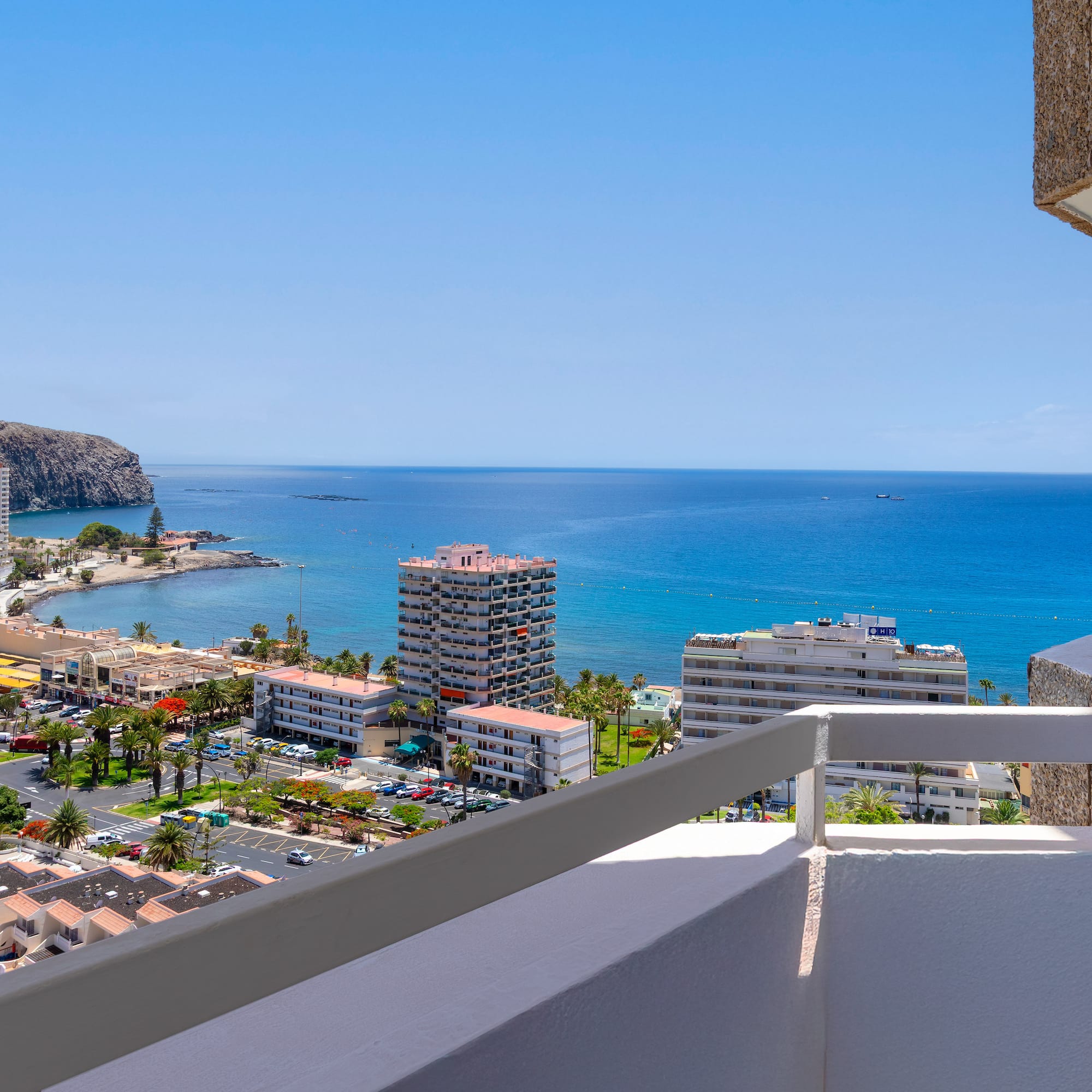 Panoramic balcony view of a sunny coastal town, blue ocean, and rugged cliffs.