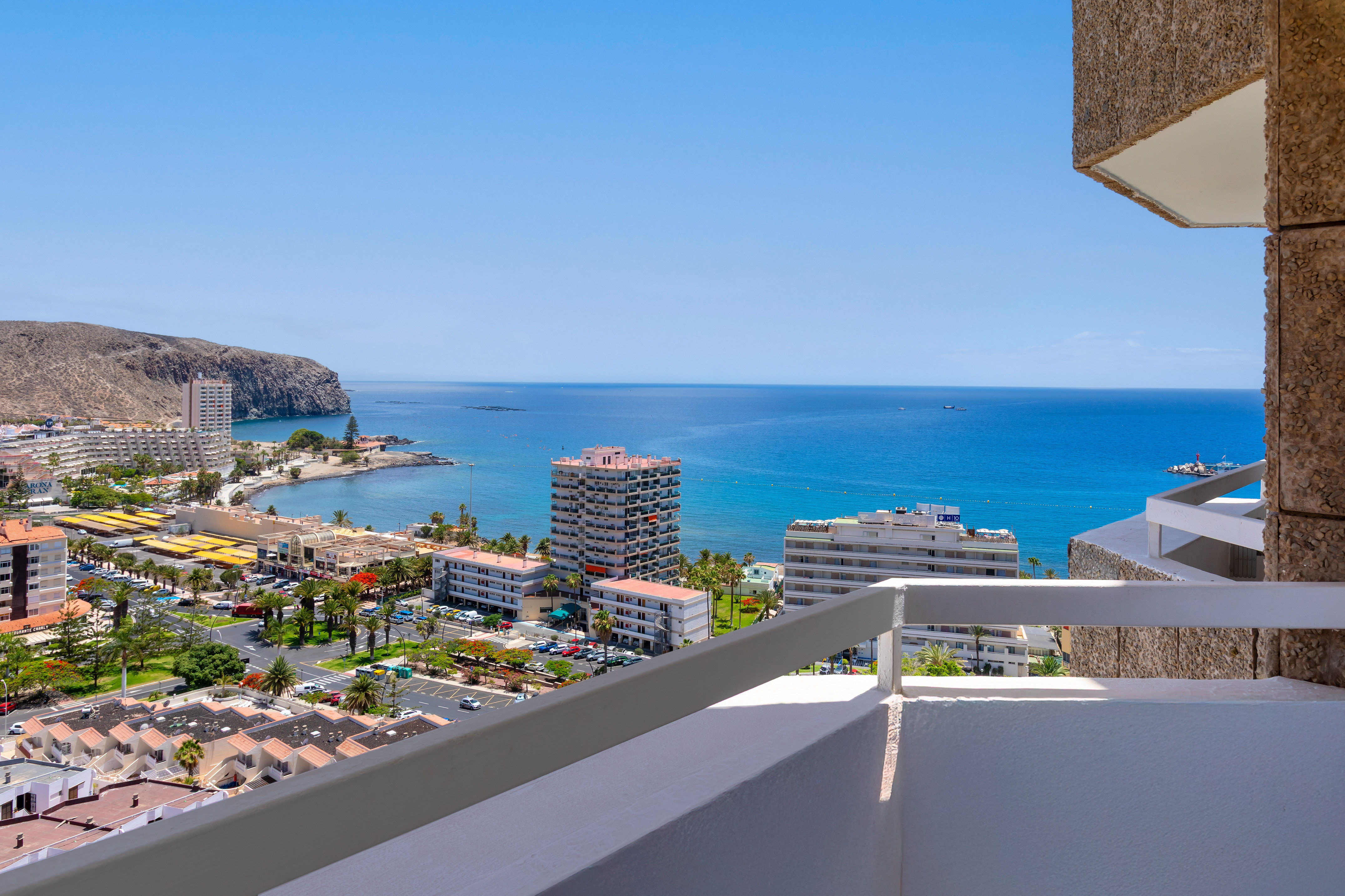 Panoramic balcony view of a sunny coastal town, blue ocean, and rugged cliffs.