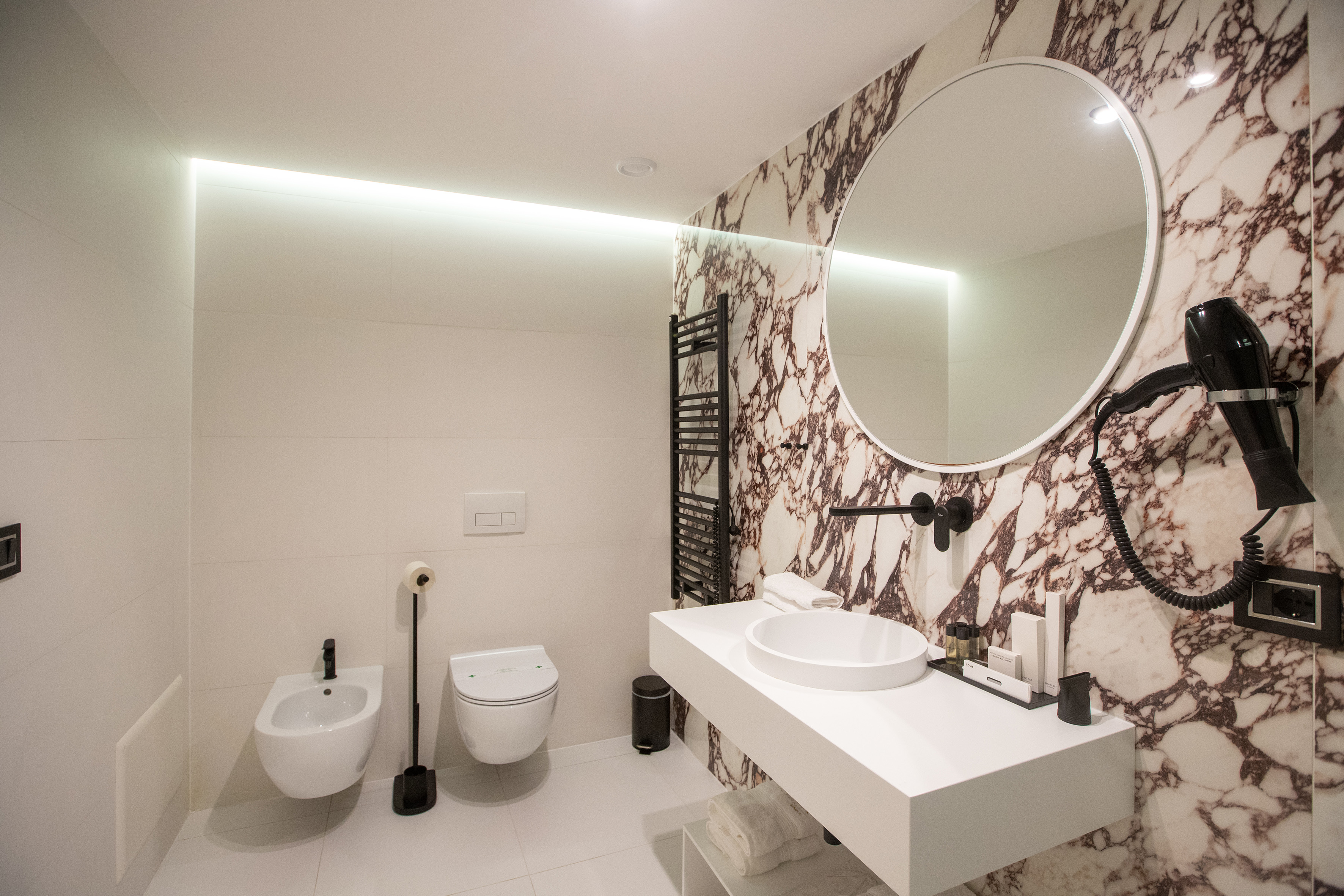Modern bathroom with marble walls, round mirror, floating vanity, toilet, and bidet.