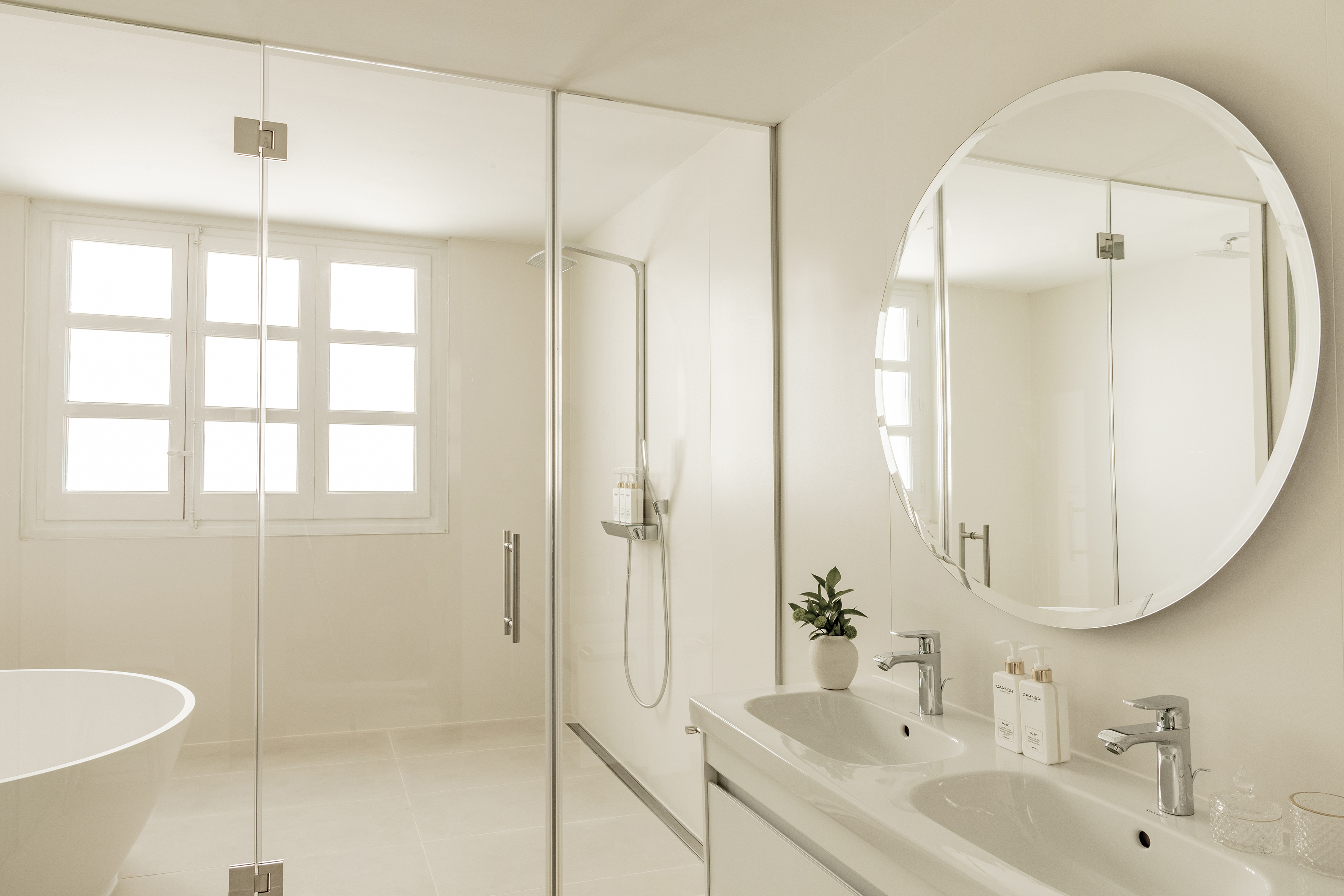 Bright minimalist bathroom with glass shower, double vanity, oval mirror.