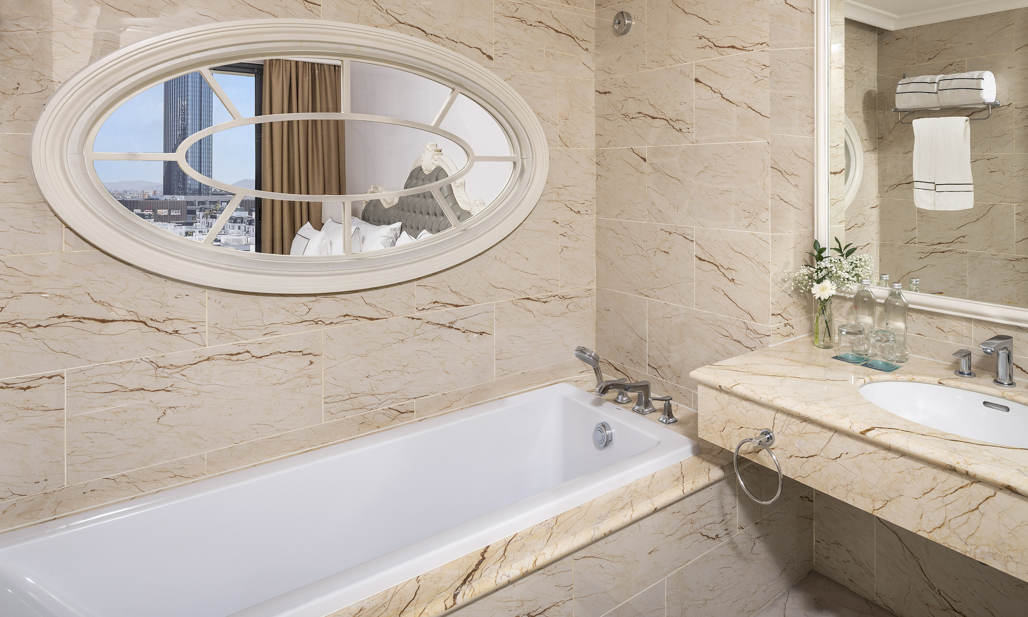 Elegant hotel bathroom with marble, bathtub, vanity, and oval mirror reflecting city view.