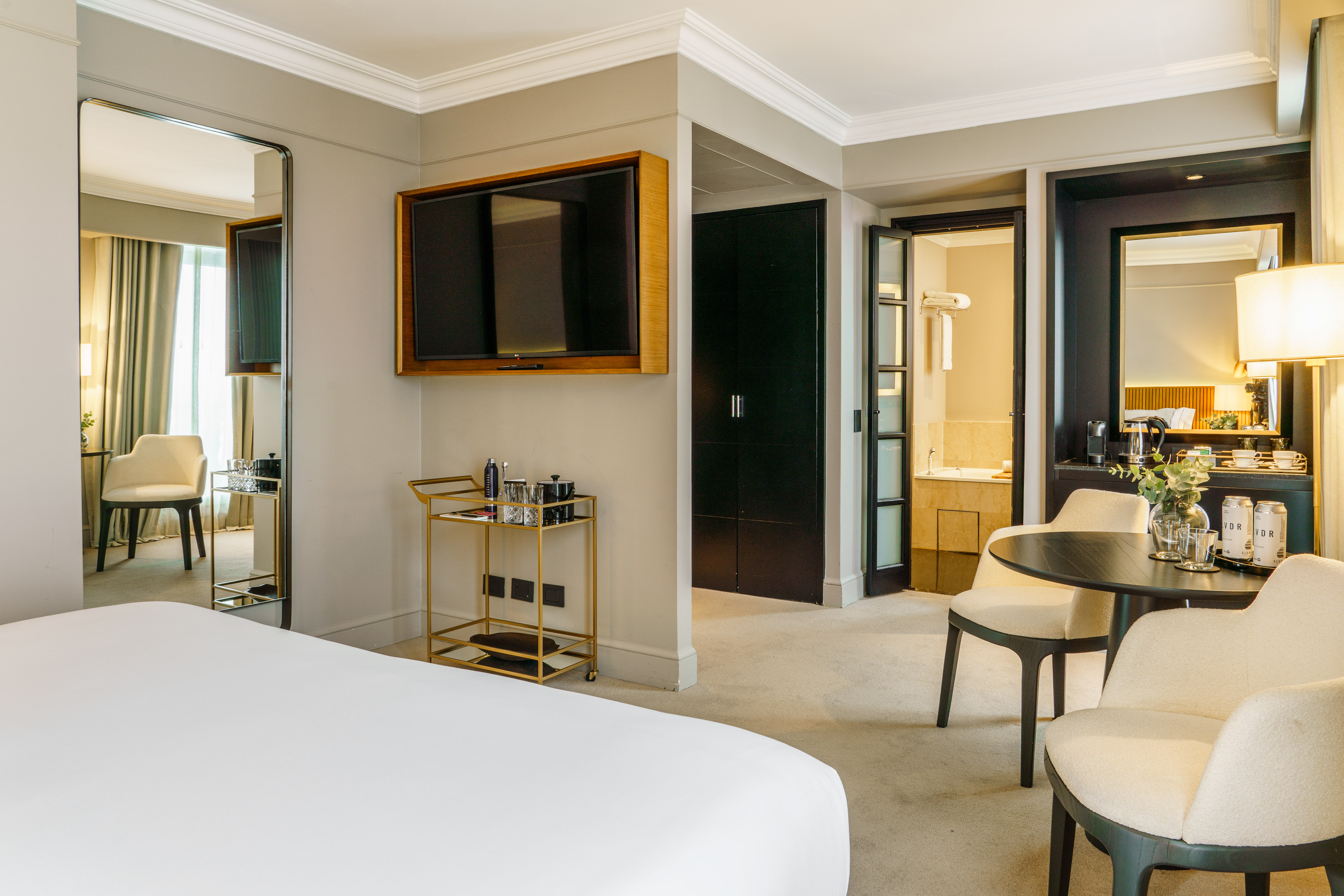 Modern luxury hotel suite with king bed, wall TV, bar cart, and open bathroom.