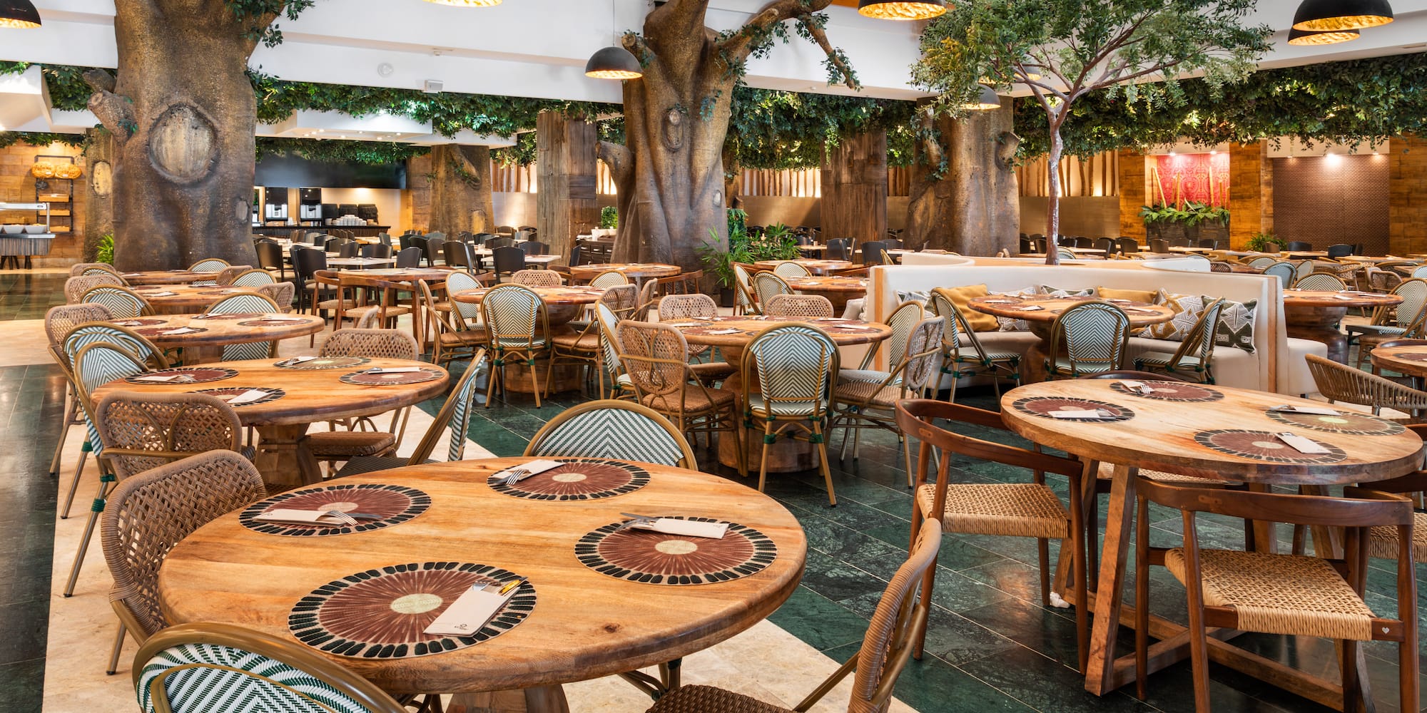 Modern restaurant with jungle decor, tree columns, wooden tables, and woven chairs.