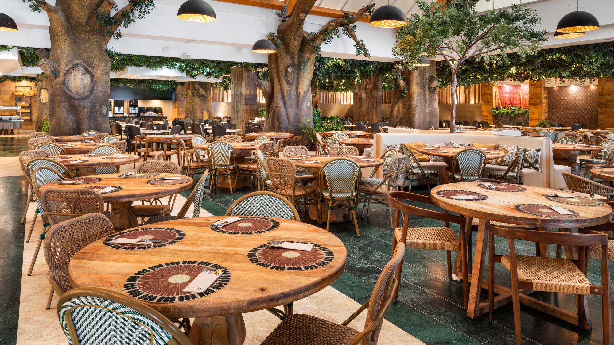 Lush forest restaurant with large trees, wooden tables, and rattan chairs.