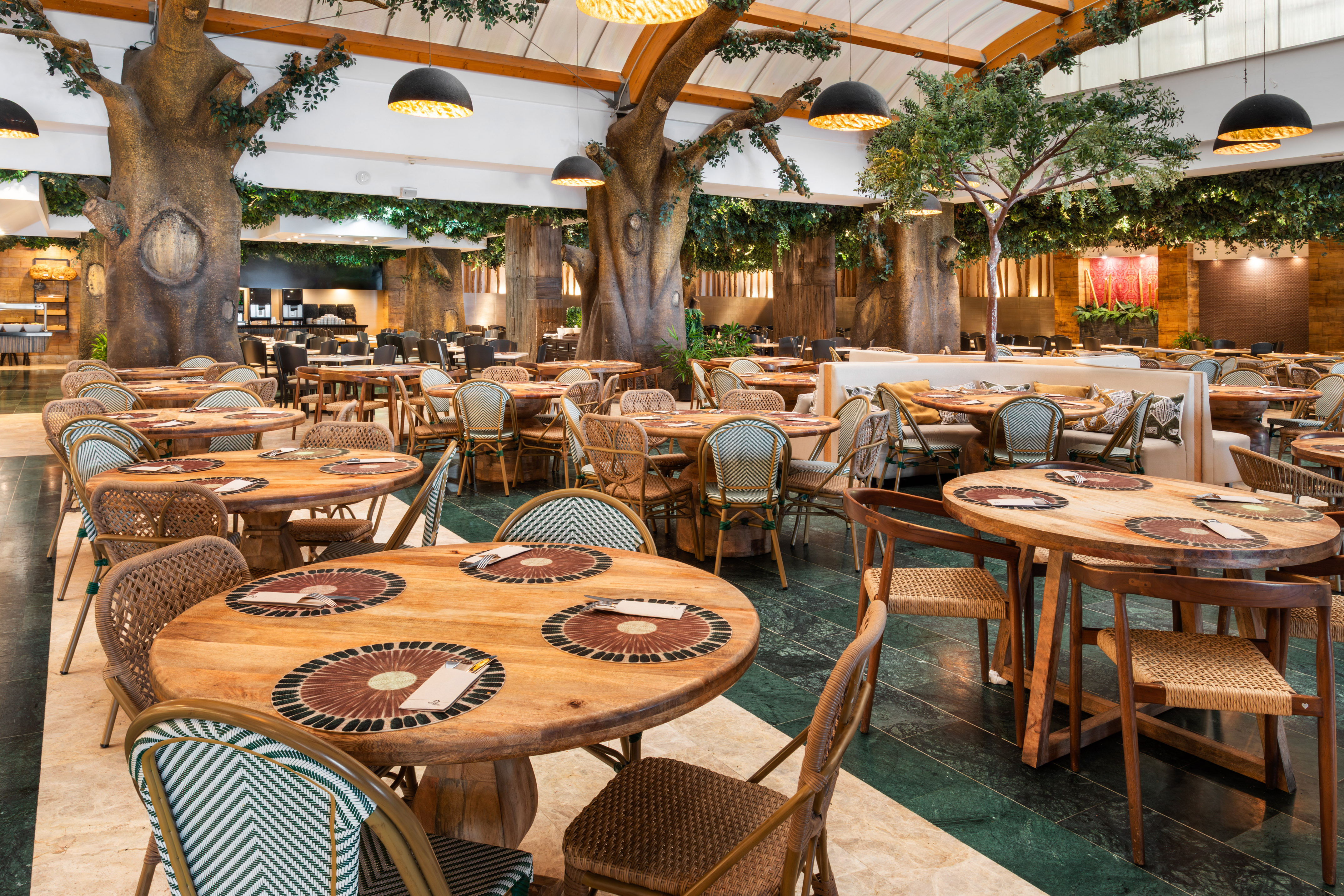 Modern restaurant with jungle decor, tree columns, wooden tables, and woven chairs.