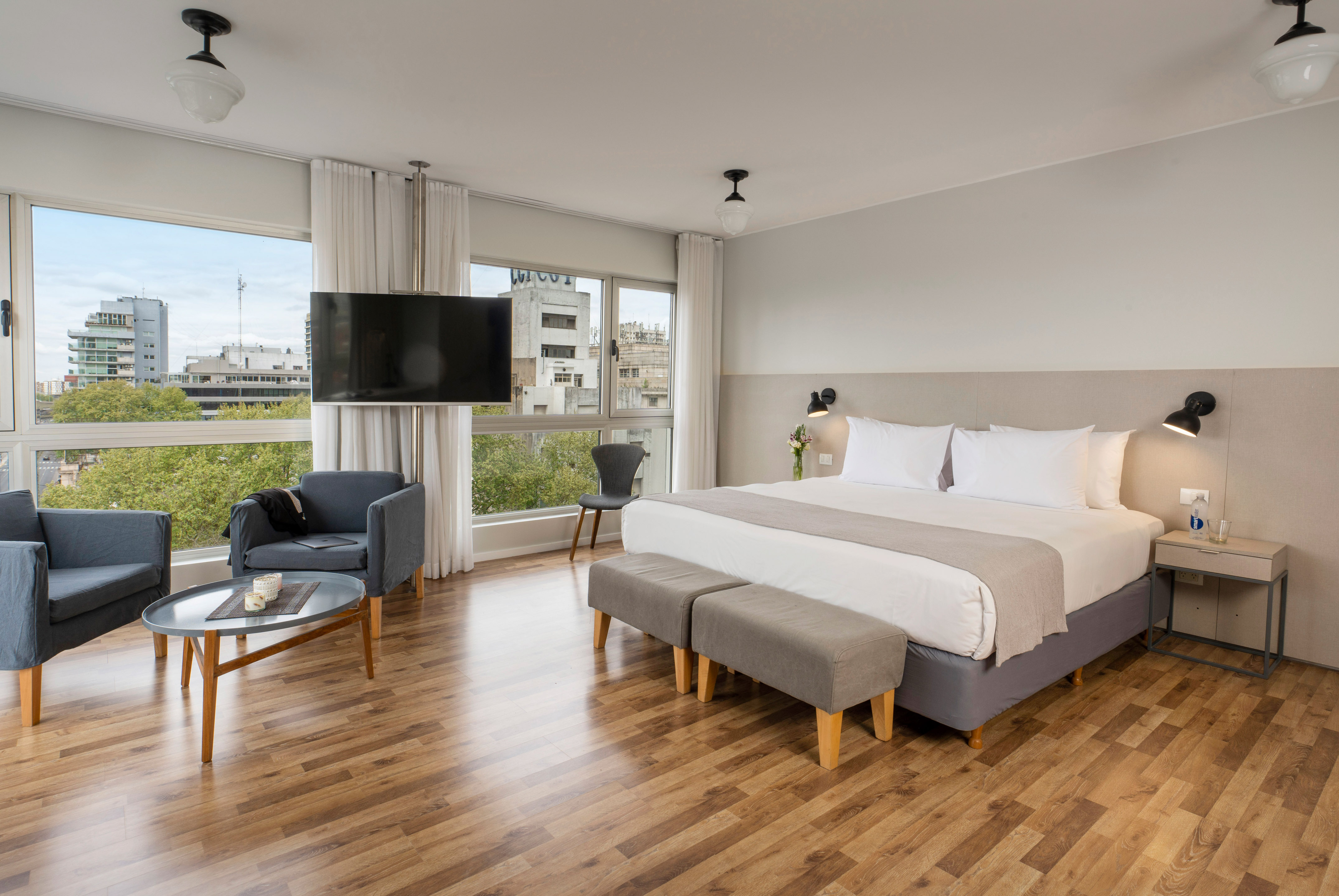 Modern hotel room with king bed, wood floor, city view, TV, and seating area.