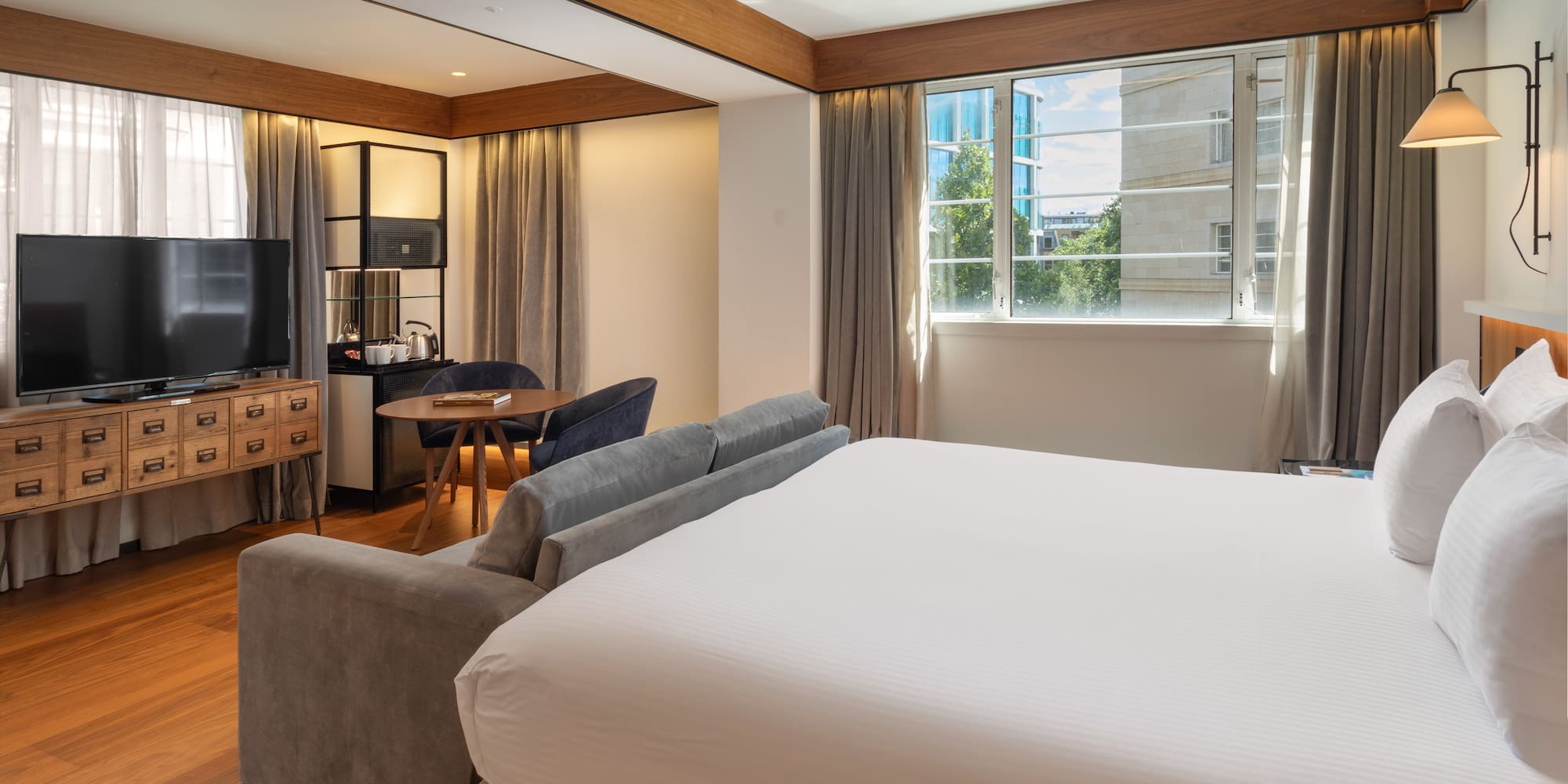 Modern hotel suite with king bed, sofa, workspace, and city view.
