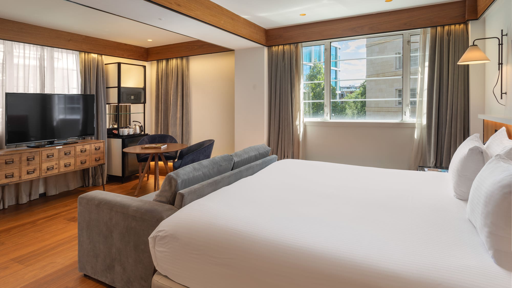 Modern hotel suite with king bed, sofa, workspace, and city view.