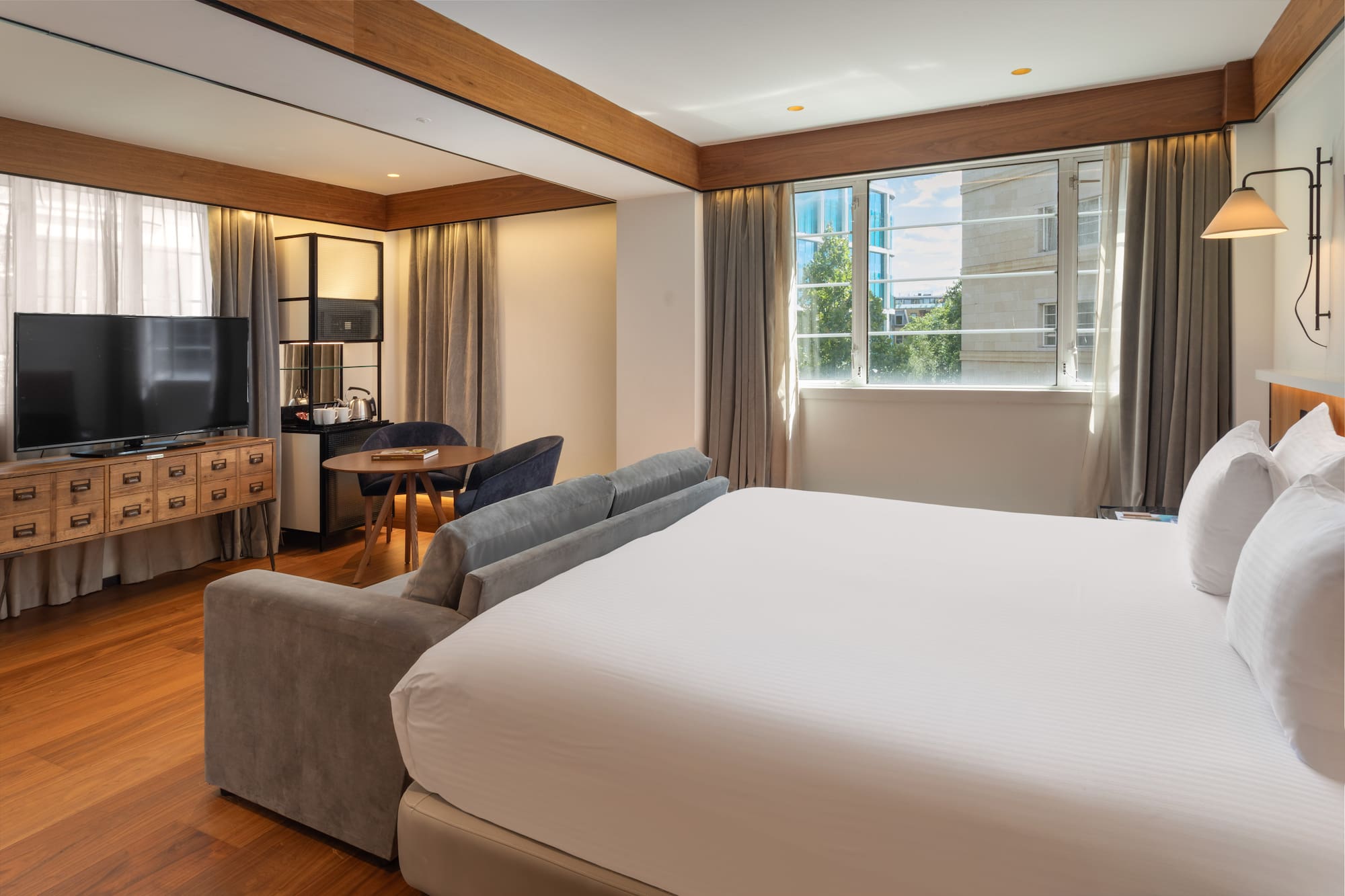 Modern hotel suite with king bed, sofa, workspace, and city view.