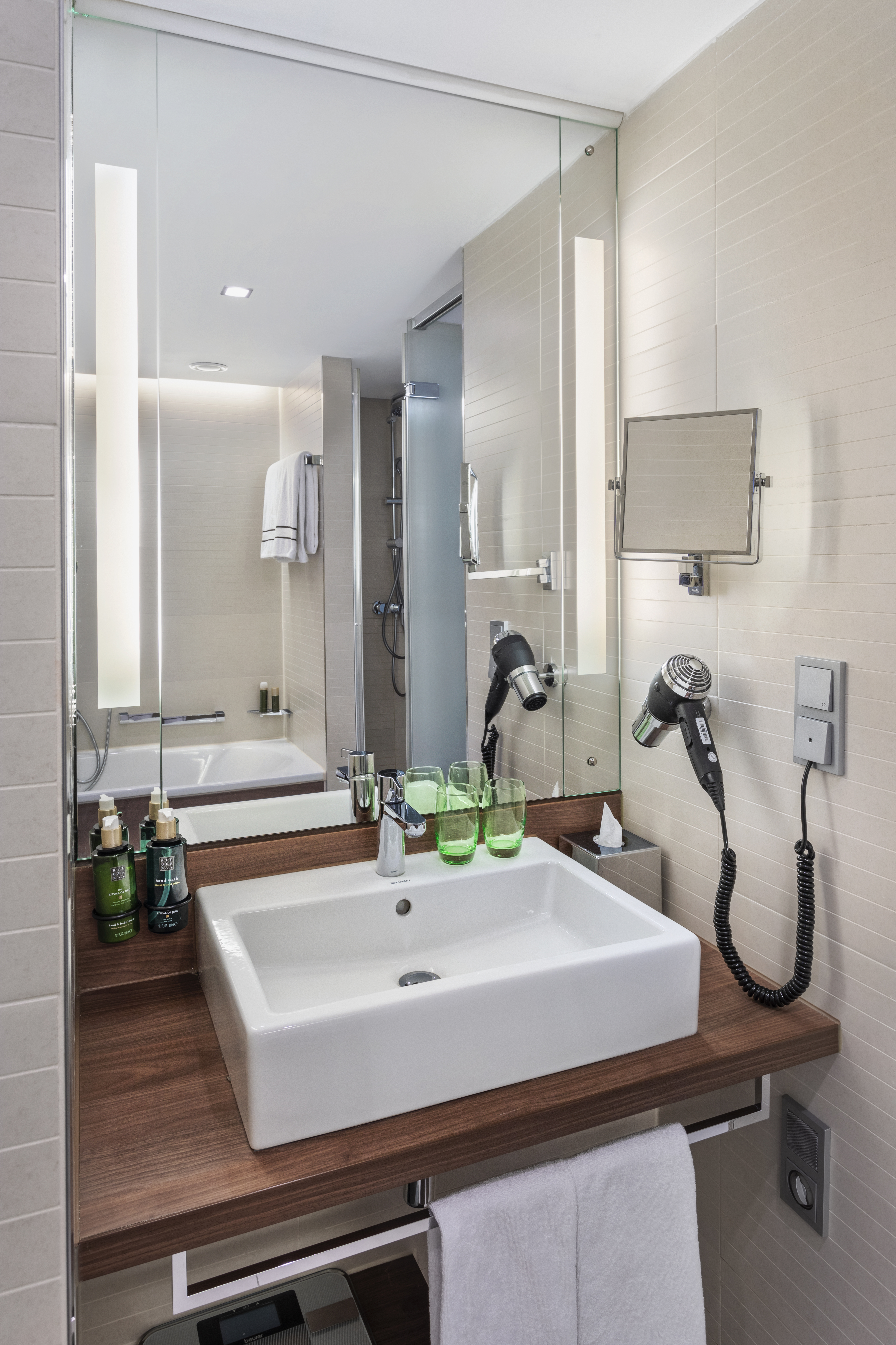 Modern hotel bathroom with white sink, wooden vanity, lighted mirror, and shower.