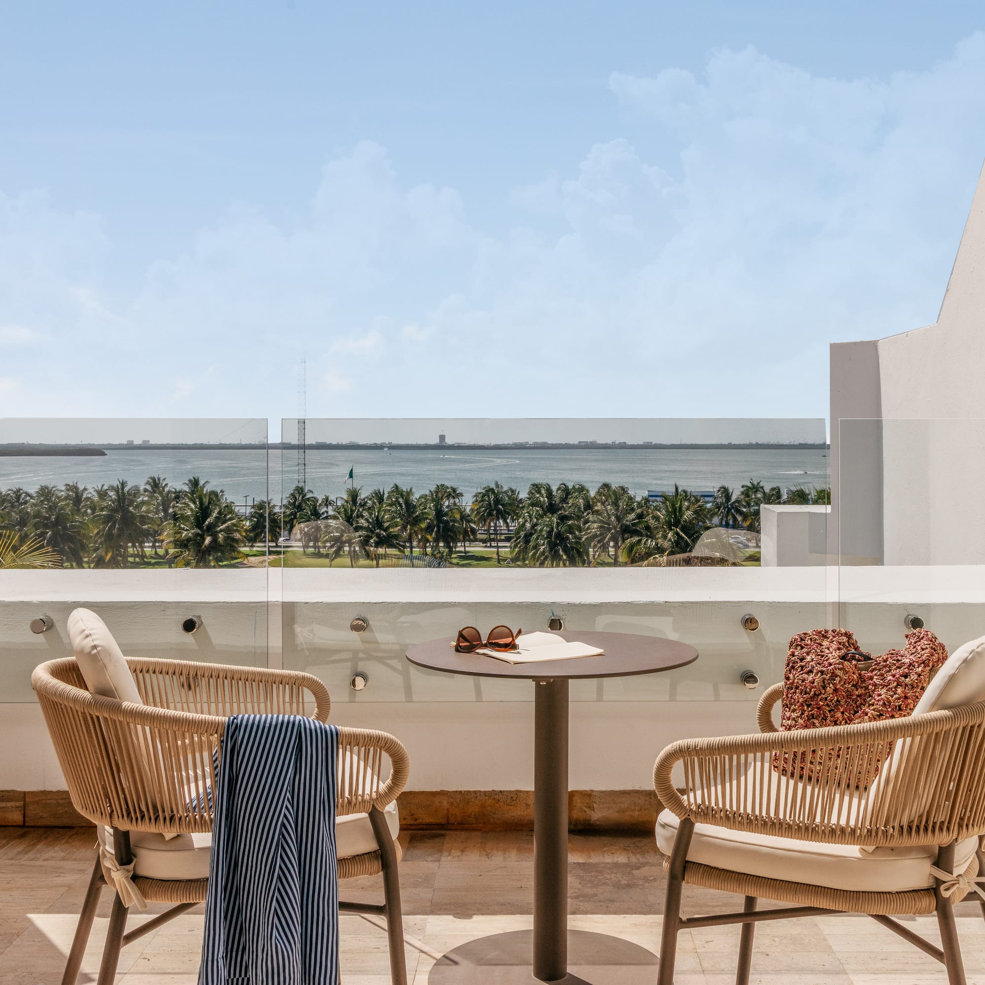 Hotel balcony with chairs, table, overlooking tropical ocean and palm trees.