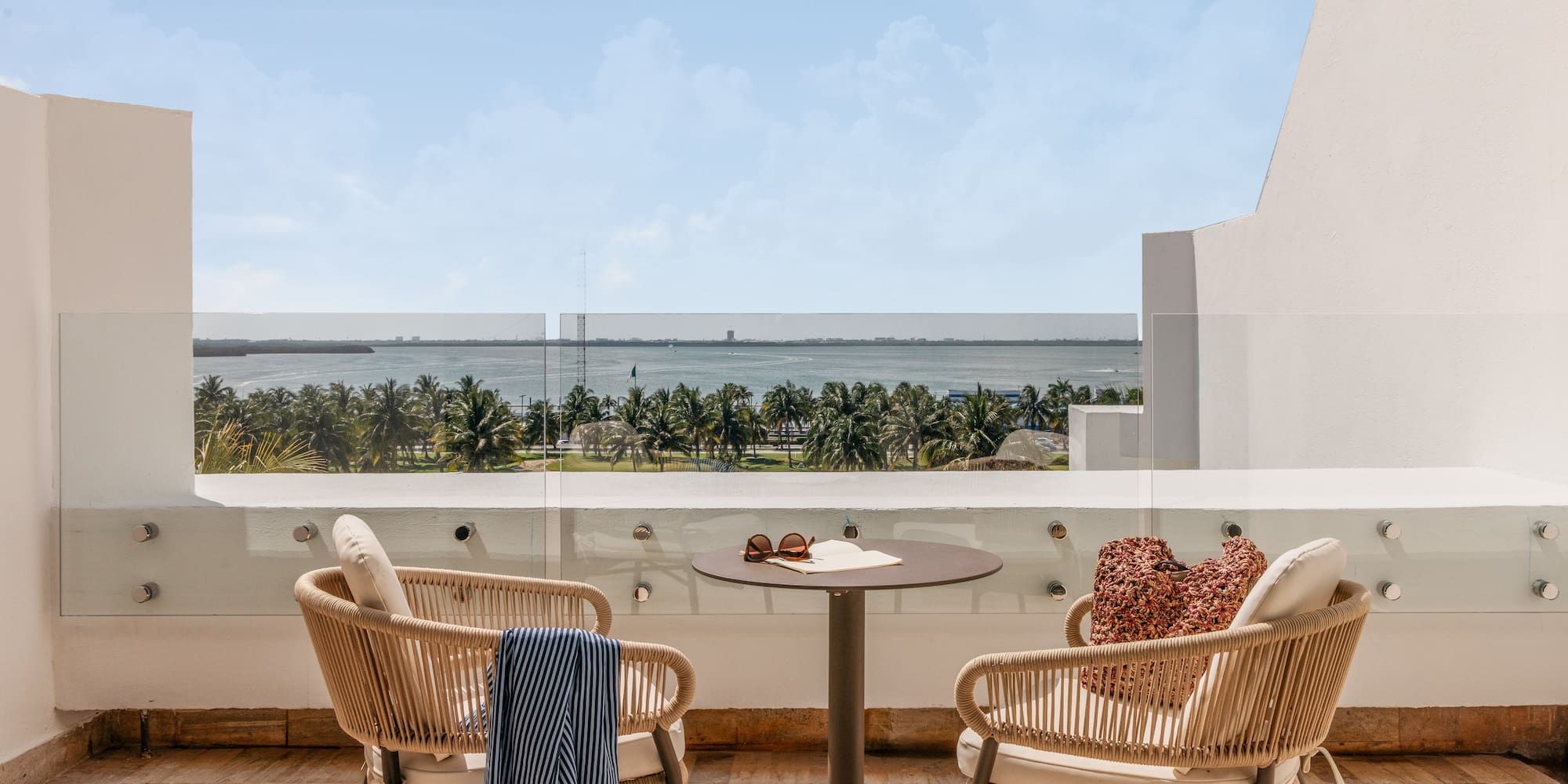 Hotel balcony with chairs, table, overlooking tropical ocean and palm trees.