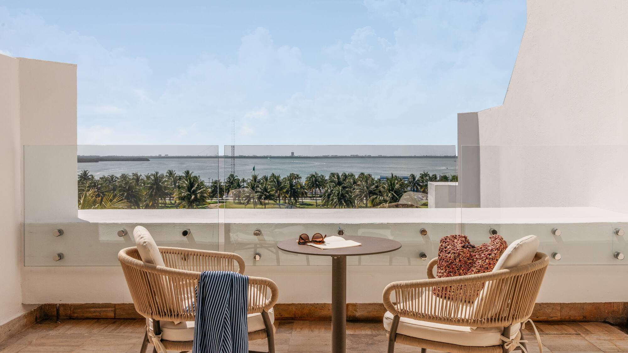 Hotel balcony with chairs, table, overlooking tropical ocean and palm trees.
