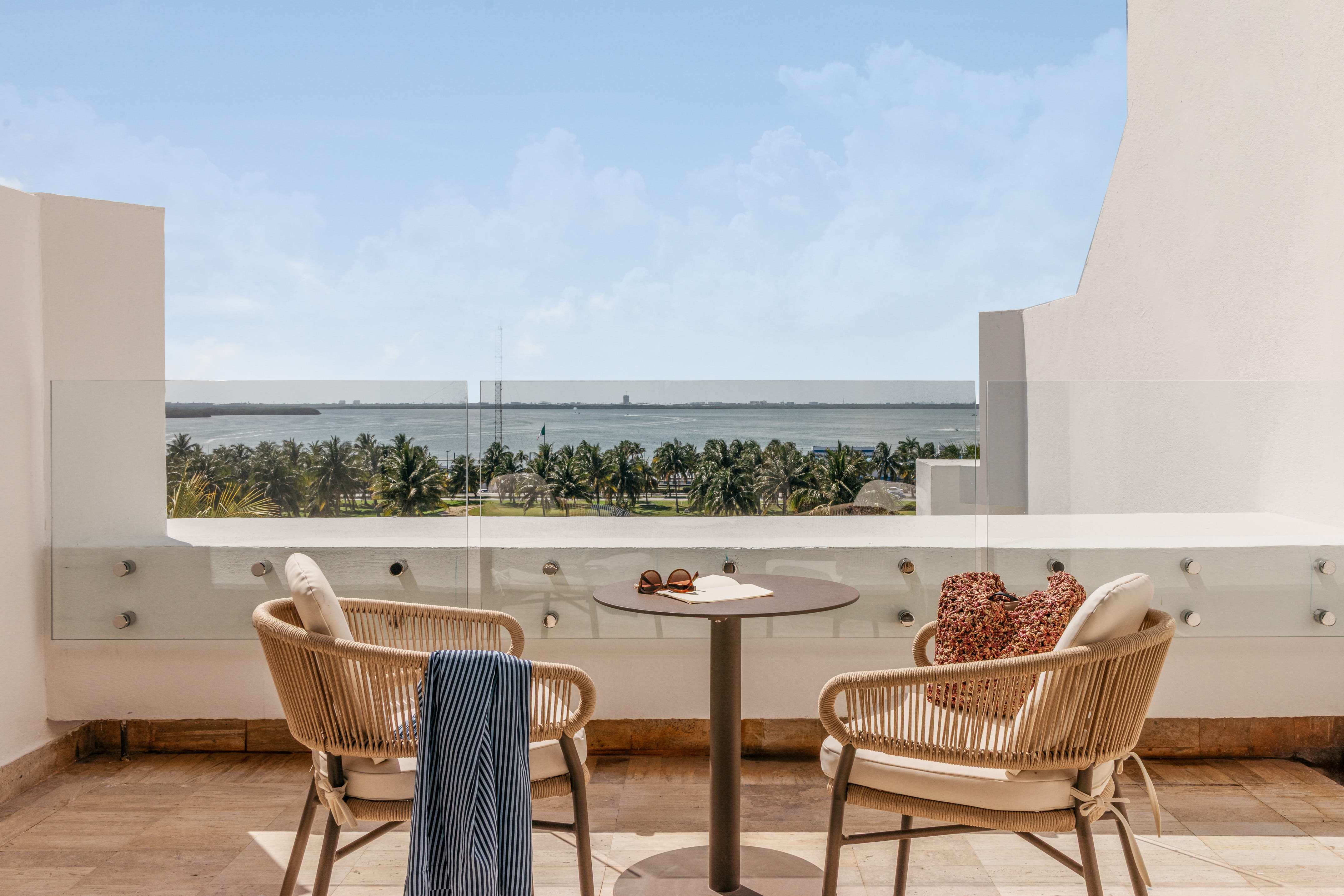 Hotel balcony with chairs, table, overlooking tropical ocean and palm trees.