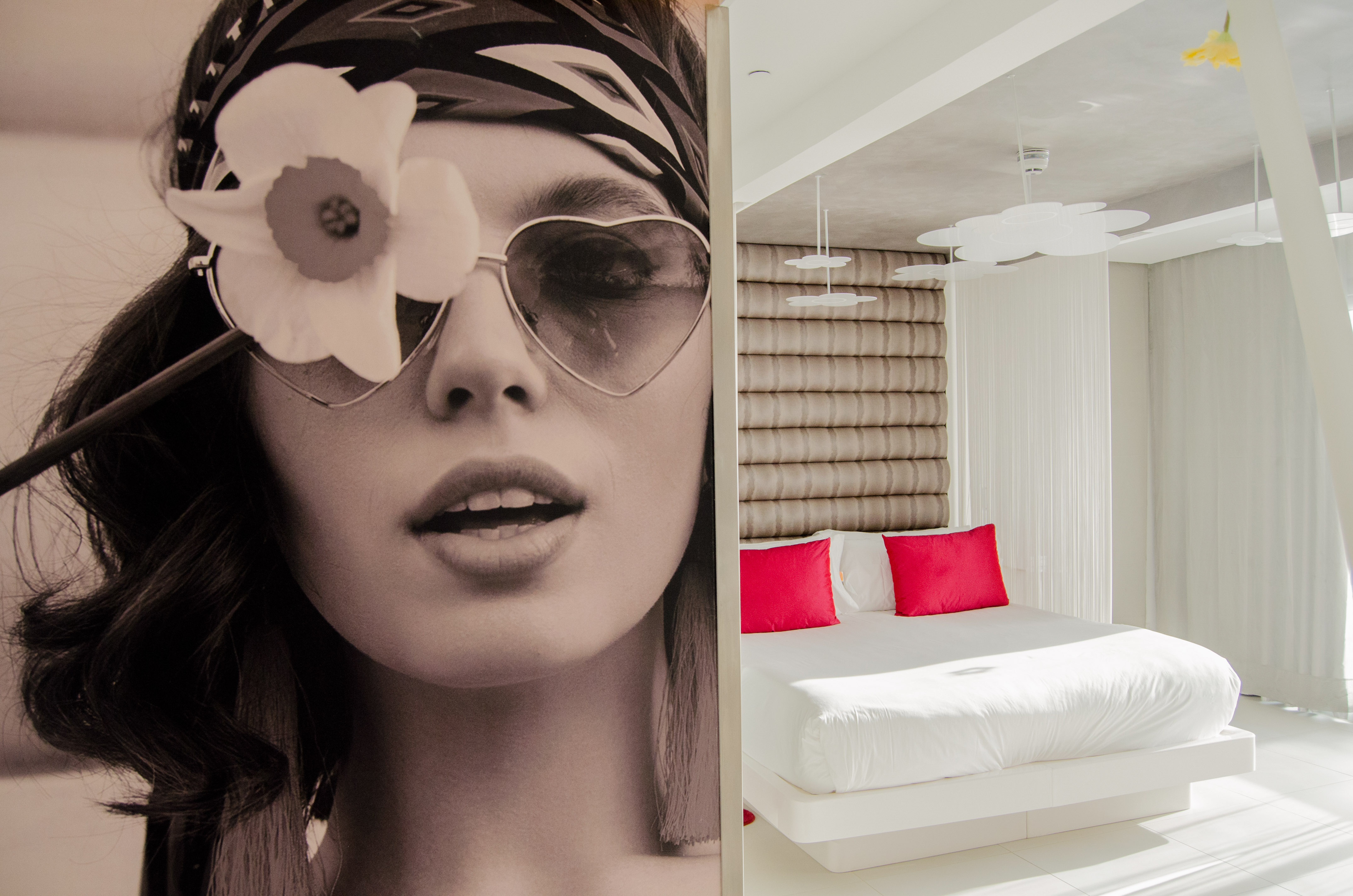 Modern hotel bedroom: white bed, red pillows, and large woman portrait wall art.