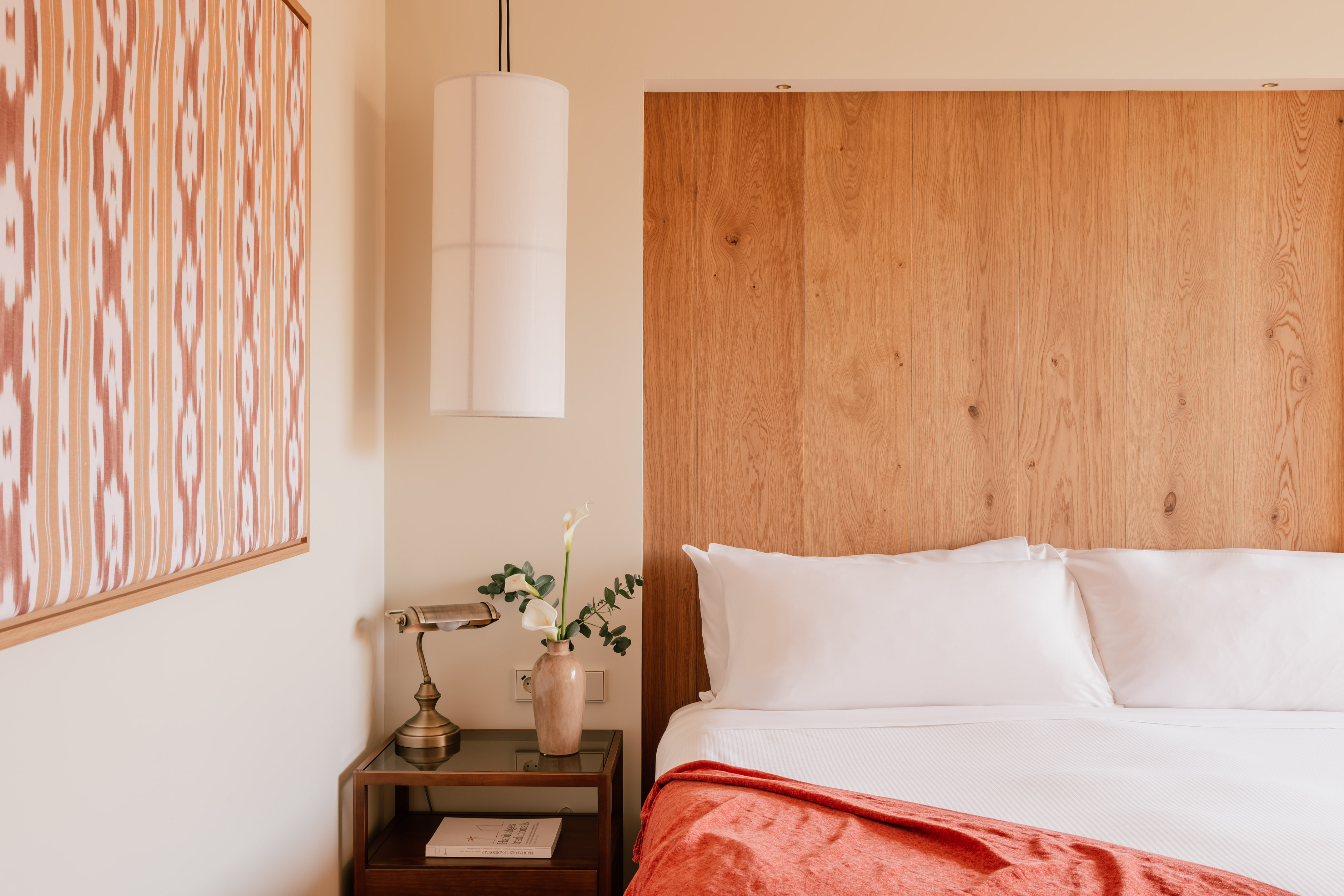 Clean hotel bedroom with wooden headboard, white bed, coral blanket, nightstand.