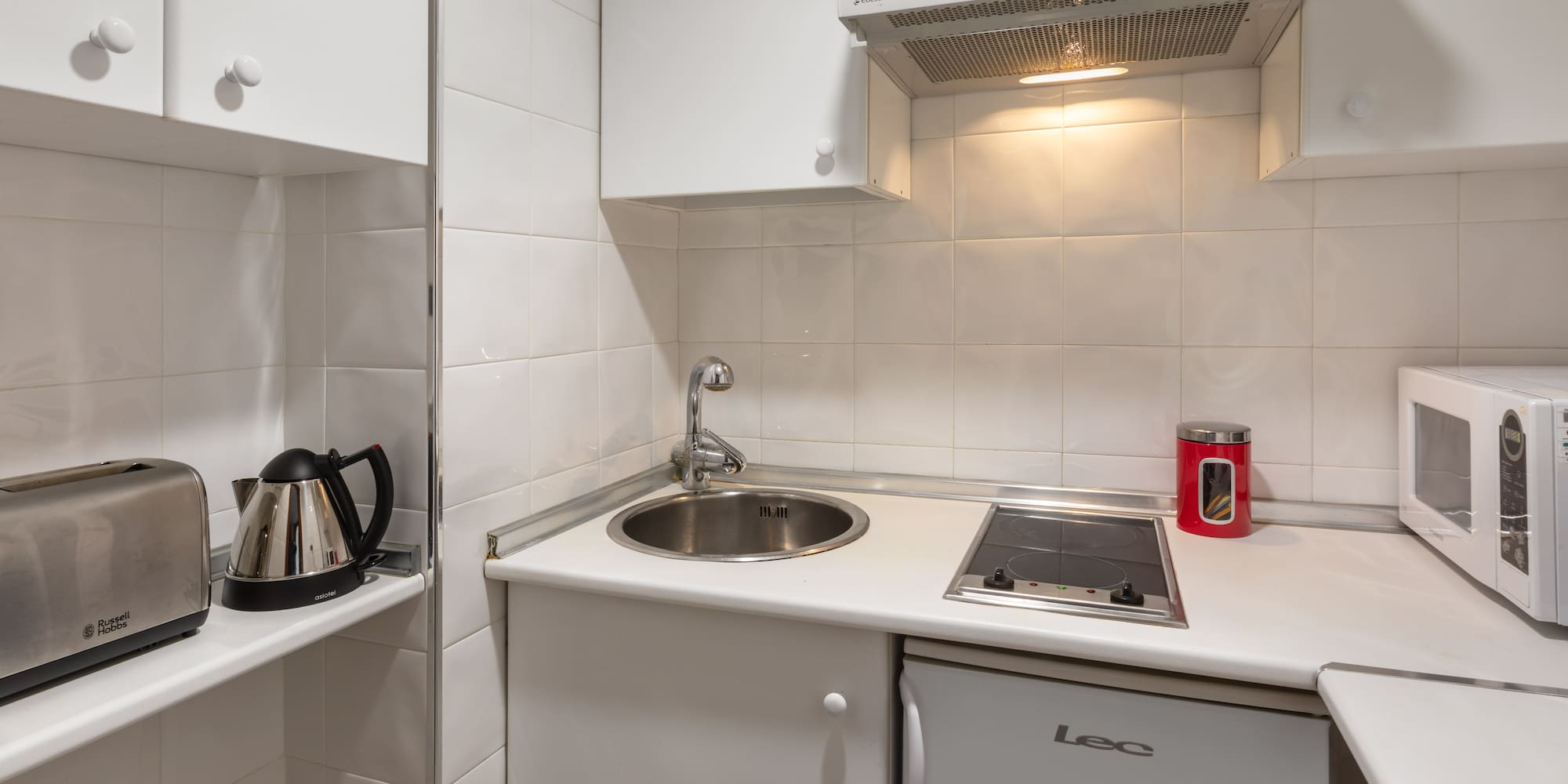 Compact white kitchenette with sink, stove, fridge, microwave, toaster, and kettle.