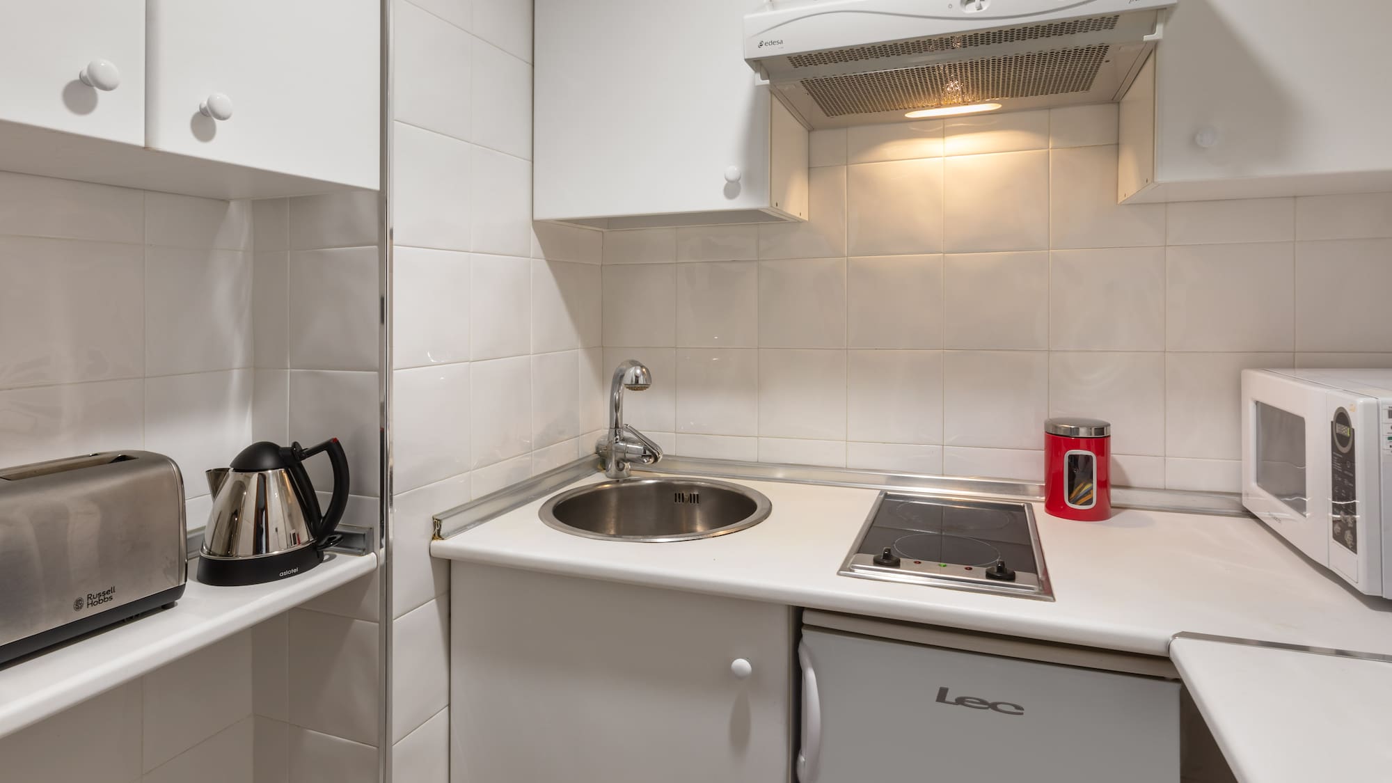Compact white kitchenette with sink, stove, fridge, microwave, toaster, and kettle.