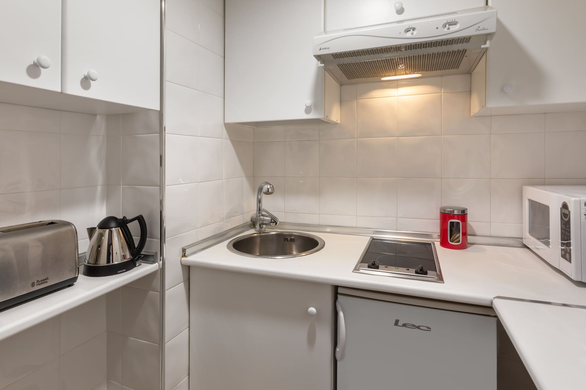 Compact white kitchenette with sink, stove, fridge, microwave, toaster, and kettle.