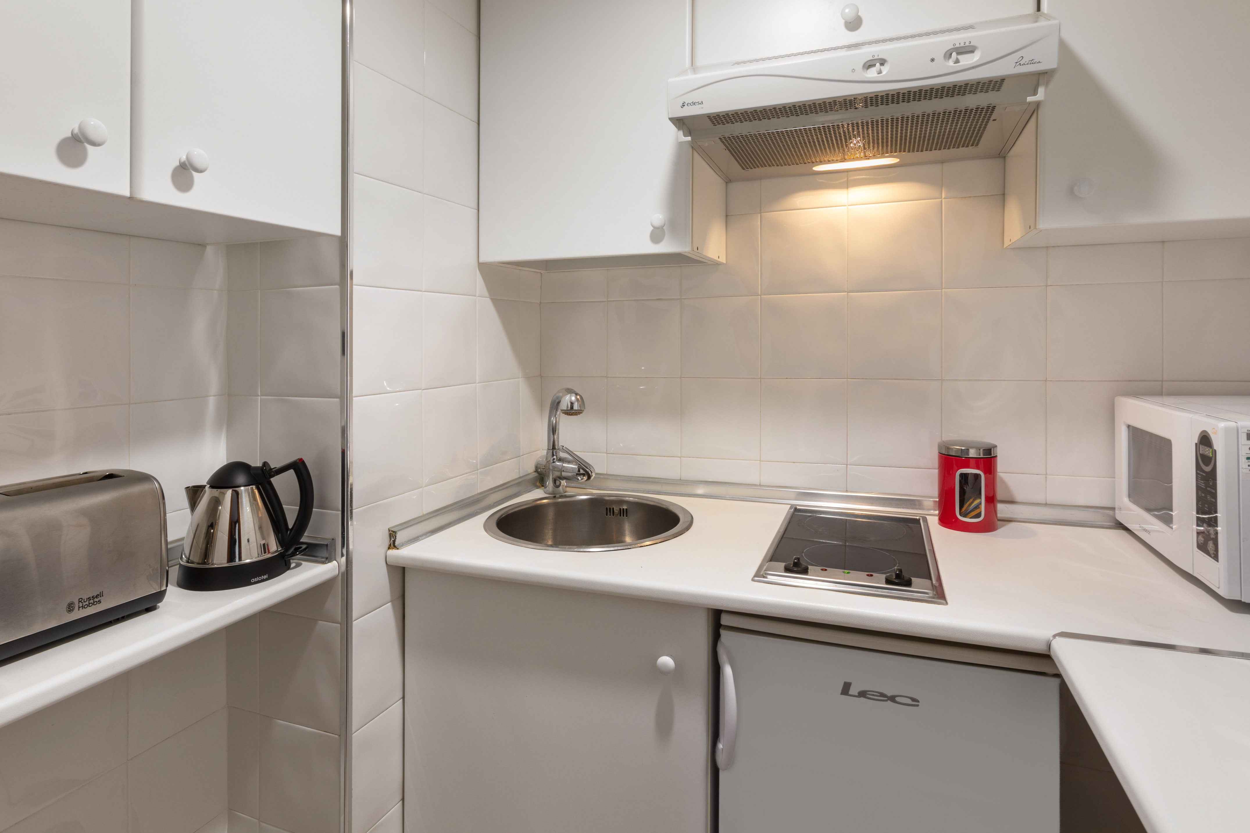 Compact white kitchenette with sink, stove, fridge, microwave, toaster, and kettle.
